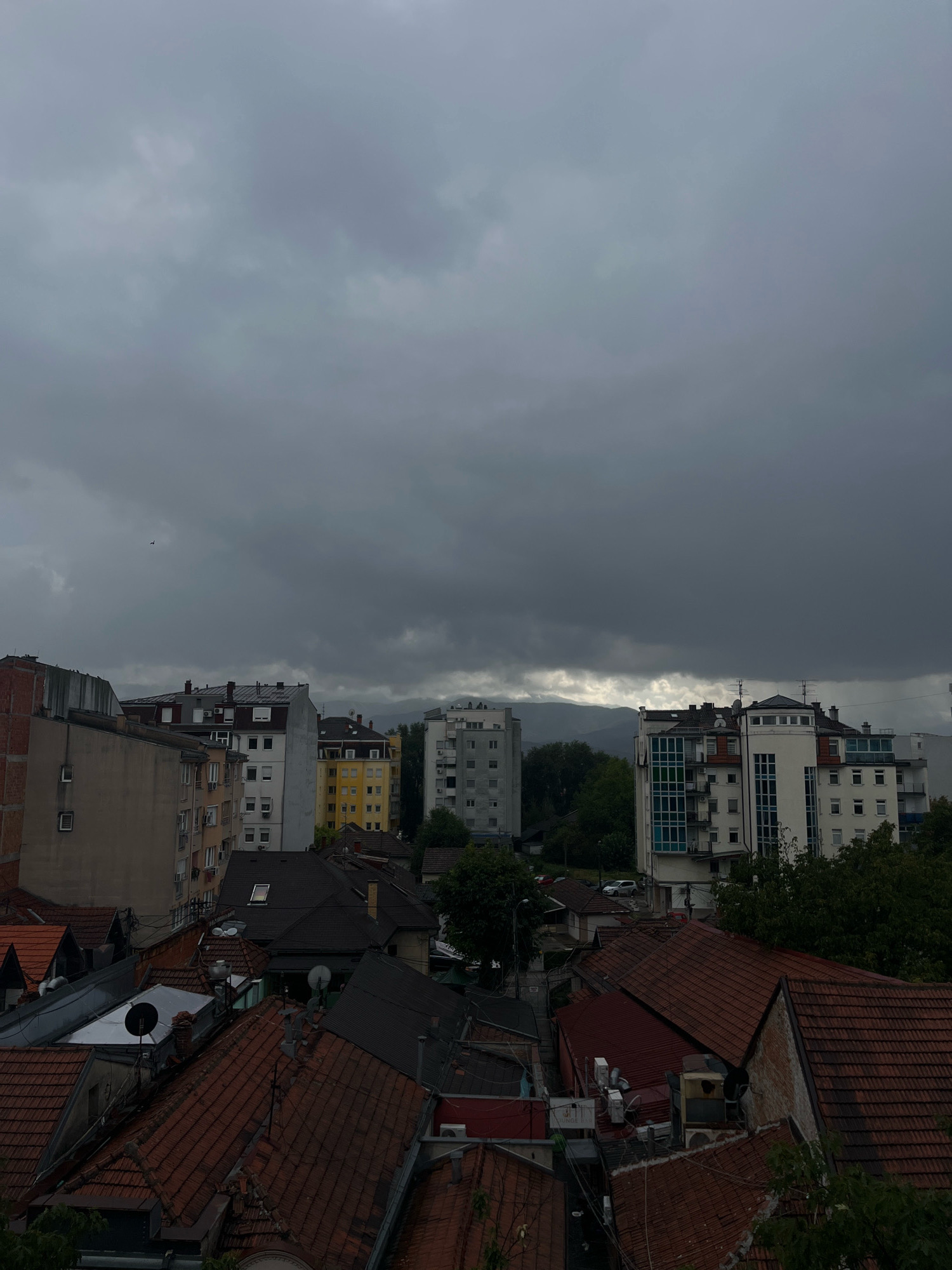 The image displays an urban landscape under a heavy, overcast sky. In the foreground, numerous red-tiled roofs of residential buildings are visible, some appearing older with visible brickwork. Behind these, apartment buildings of varying heights and architectural styles dominate the mid-ground. The buildings are predominantly in muted colors such as beige, yellow, gray, and white, with some featuring prominent window structures and balconies. Trees, some with dark green foliage, are interspersed among the buildings. In the far background, mountains are faintly discernible through the thick cloud cover. The overall atmosphere is somber, suggesting an impending or ongoing rain. The scene is likely located in the city of Kraljevo, Serbia, given the context of its creation. There are no discernible people or animals in the frame. The sky is a dominant feature, filled with dark, textured clouds.