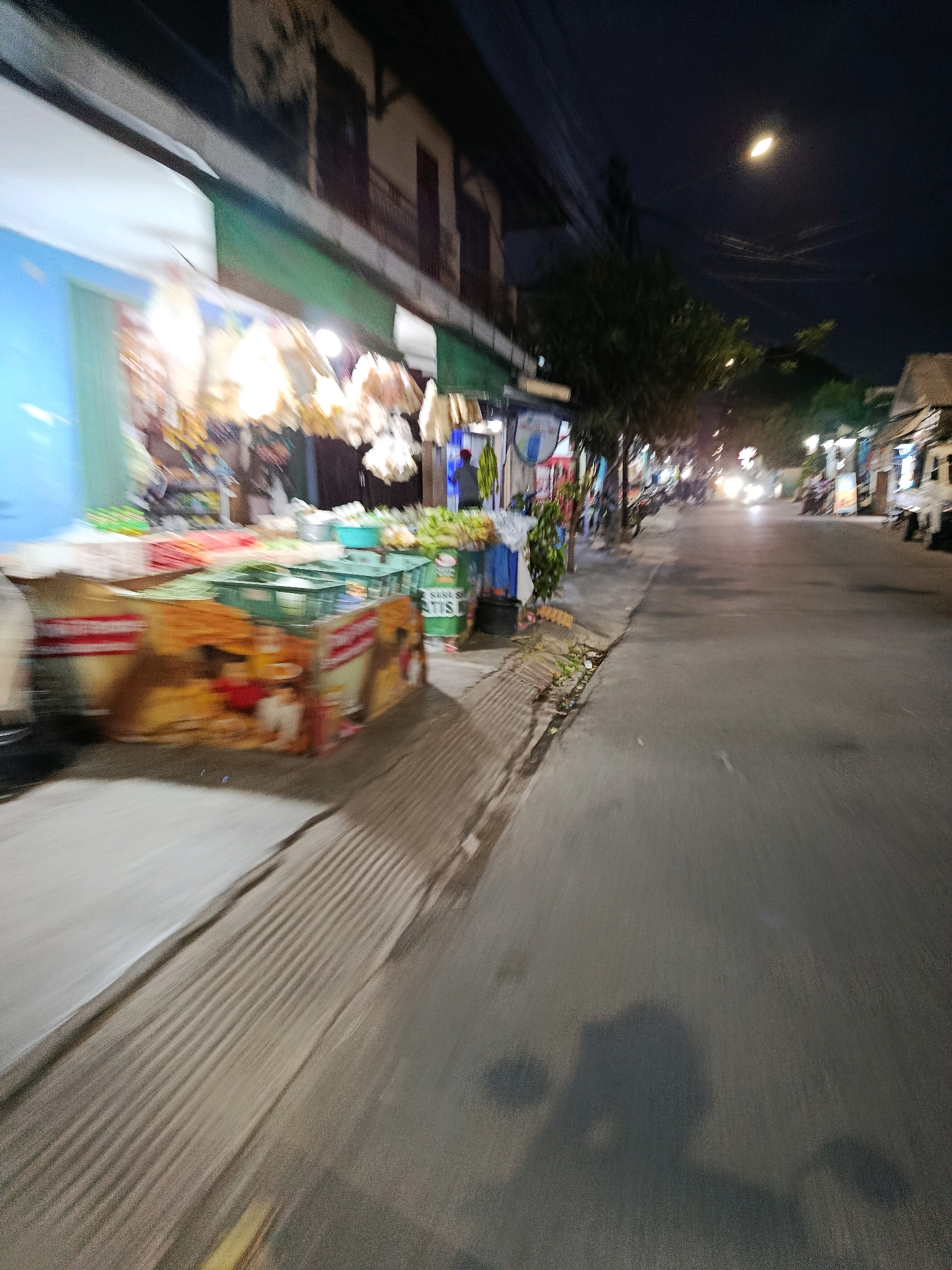 A night view shows a street market stall on the left with produce displayed, some of which is blurry due to motion. The stall is illuminated by lights and has various items for sale. The street extends into the distance with other stalls and buildings visible on both sides. Vehicles are seen on the street, with their headlights on. The overall scene suggests a lively market atmosphere at night in Bekasi, Indonesia.