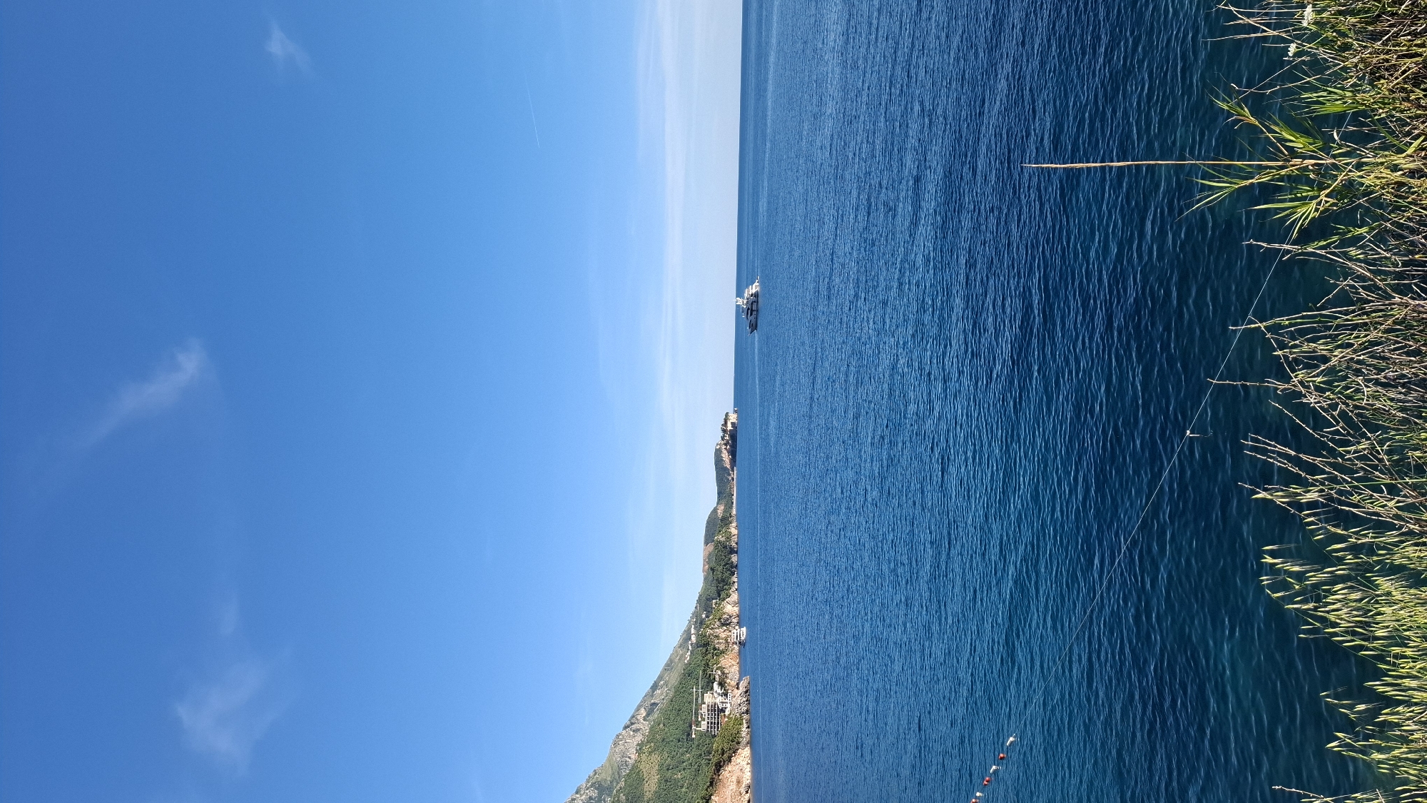 The image presents a seascape under a clear blue sky with scattered wispy clouds. A portion of the landmass is visible, with buildings nestled along the coastline. A ship is stationed on the water. Tall grass and vegetation occupy the right edge of the frame. The location appears to be coastal, but the specific city or region is unknown.