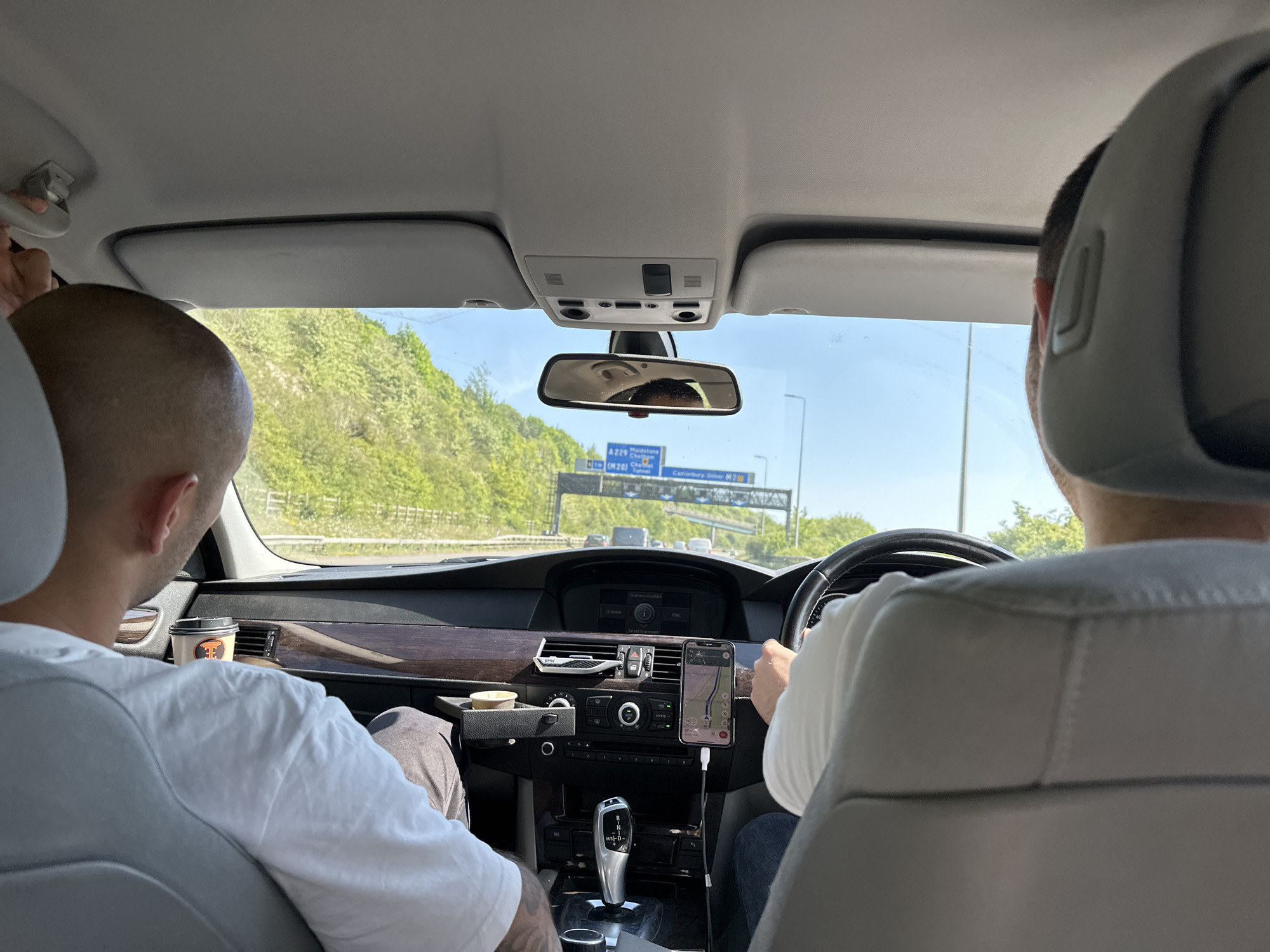 The image shows the interior of a car driving on a highway. There are two men visible: one in the passenger seat and one driving. The driver has their hands on the steering wheel, and a smartphone displaying a map is mounted on the dashboard. A road sign visible in the windshield indicates directions to "A229 Maidstone, (M20) Tunnel, Canterbury, Dover (M2)." Coffee cups are visible in the car. The landscape outside the window includes green trees and a clear sky. The location is Unknown, Unknown.