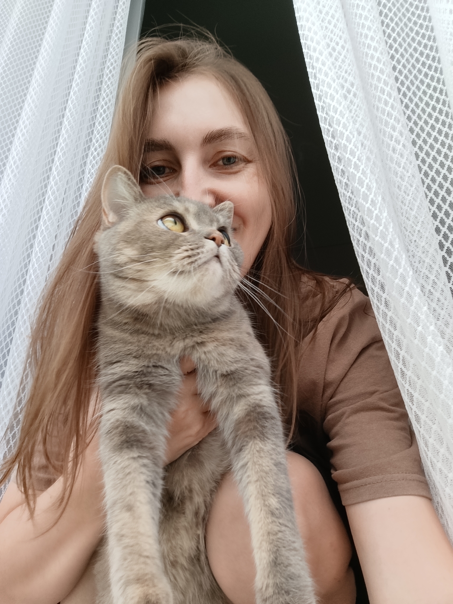 In a selfie, a young woman with long, light brown hair and light blue eyes is holding a grey tabby cat. The woman is smiling as she looks up and to the left, with the cat looking in the same direction. They are positioned in front of a sheer white curtain. The background is dark, suggesting an indoor setting. The image was taken at the intersection of Mira Street and Karl Marx Street in Chaikovsky, Russia.