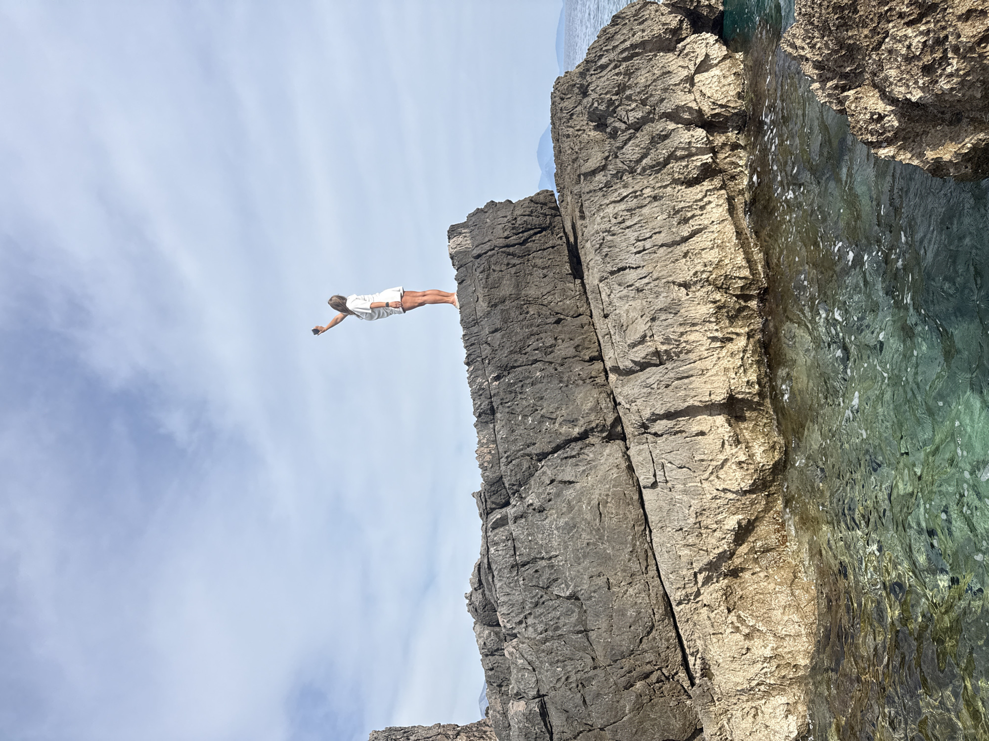 A woman is poised on a rocky cliff edge on a sunny day. She is wearing a white shirt and shorts, her body is parallel to the ground, arms extended. Below the cliff, clear water meets the rock face. The sky is blue with scattered clouds. The location is likely Seoca, Montenegro, inferred from the image's provenance and the terrain visible.