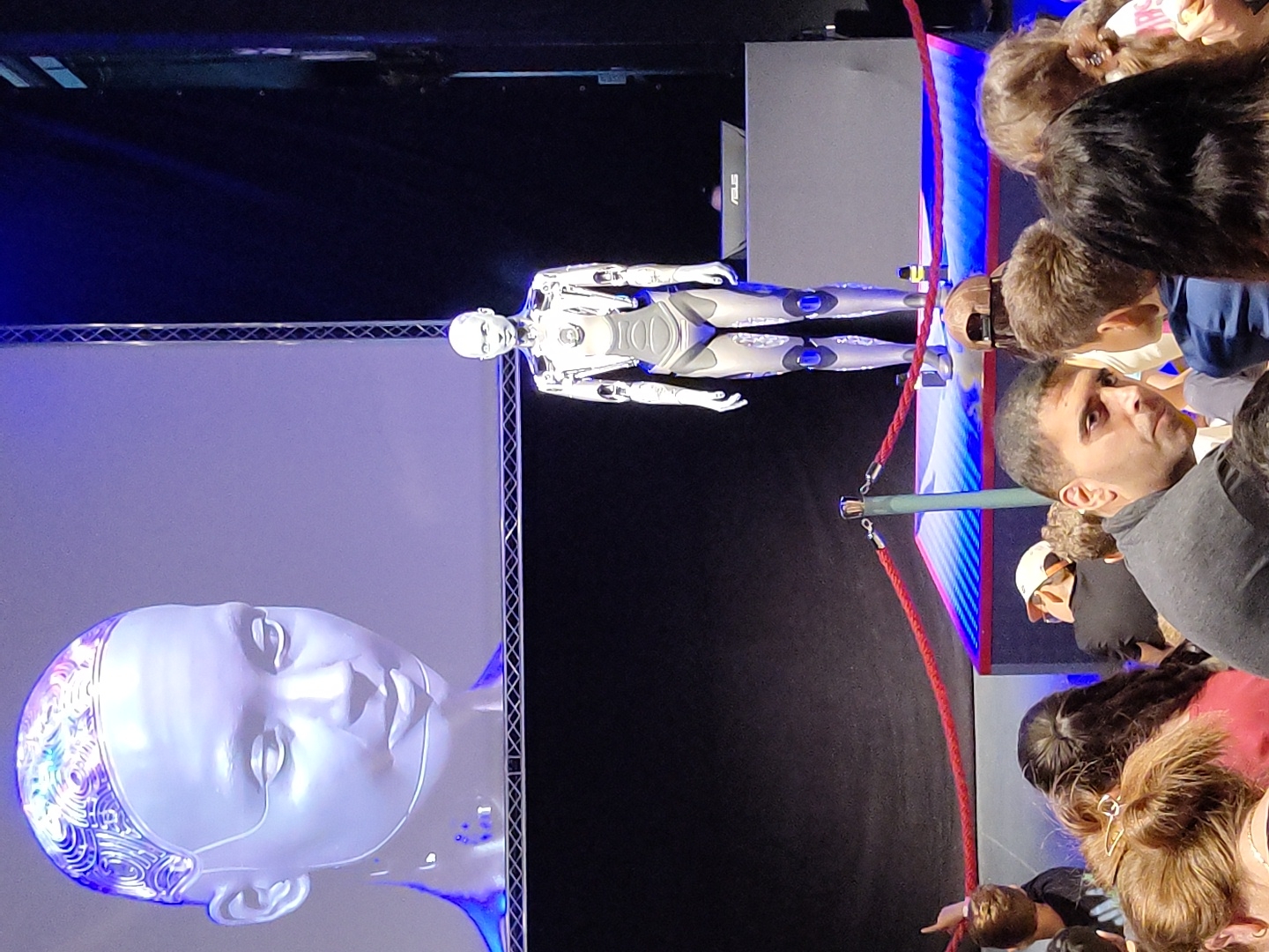 The image depicts an event featuring robots and a large audience. A life-size humanoid robot stands on a platform, while a giant projection of a robot's head is displayed on a screen behind it. The platform is illuminated with blue and pink lights, and a red rope barrier separates the platform from the audience. The audience members, appearing to be a mix of adults and children, are seated or standing in the foreground, facing the stage. Some audience members are looking towards the stage, while others are looking away or at their phones. The environment appears to be indoors, likely a conference or exhibition hall. The scene is set in Haifa, Israel.