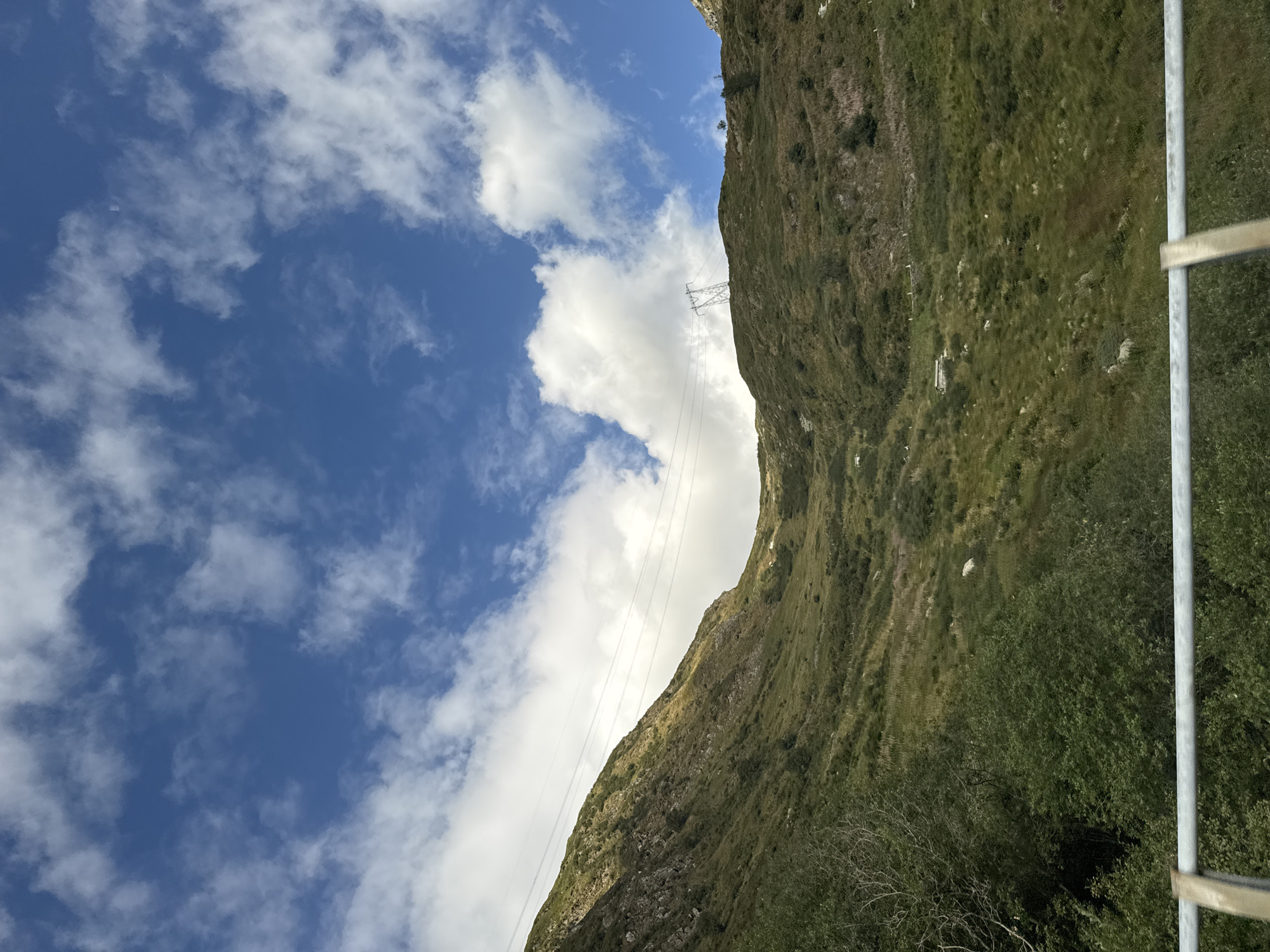 The image shows a steep, grassy mountainside under a bright blue sky dotted with white clouds. Power lines traverse the slope, supported by a tall metal tower visible on the crest of the hill. The vegetation is a mix of green grass, some rocky outcrops, and scattered bushes. In the foreground, a metal railing is visible, suggesting the vantage point is from a road or pathway. The scene appears to be a natural, mountainous landscape, possibly in the vicinity of Airolo, Switzerland, given the presence of infrastructure like power lines and the characteristic alpine terrain.