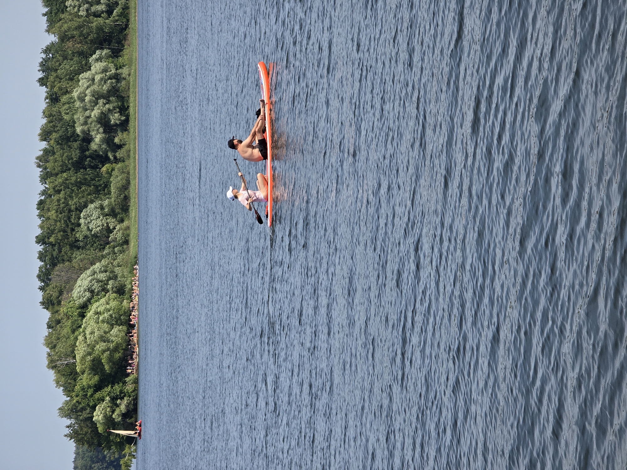 Two people are on an orange stand-up paddleboard in a body of water. The person on the left, wearing a pink shirt and white hat, is sitting and holding a paddle. The person on the right, wearing black shorts, is also sitting. They are in the foreground of the image. In the background, across the water, is a tree-lined shore with a beach where many people are gathered. A small sailboat with a tan sail is visible to the left of the shore. The sky is clear and blue.