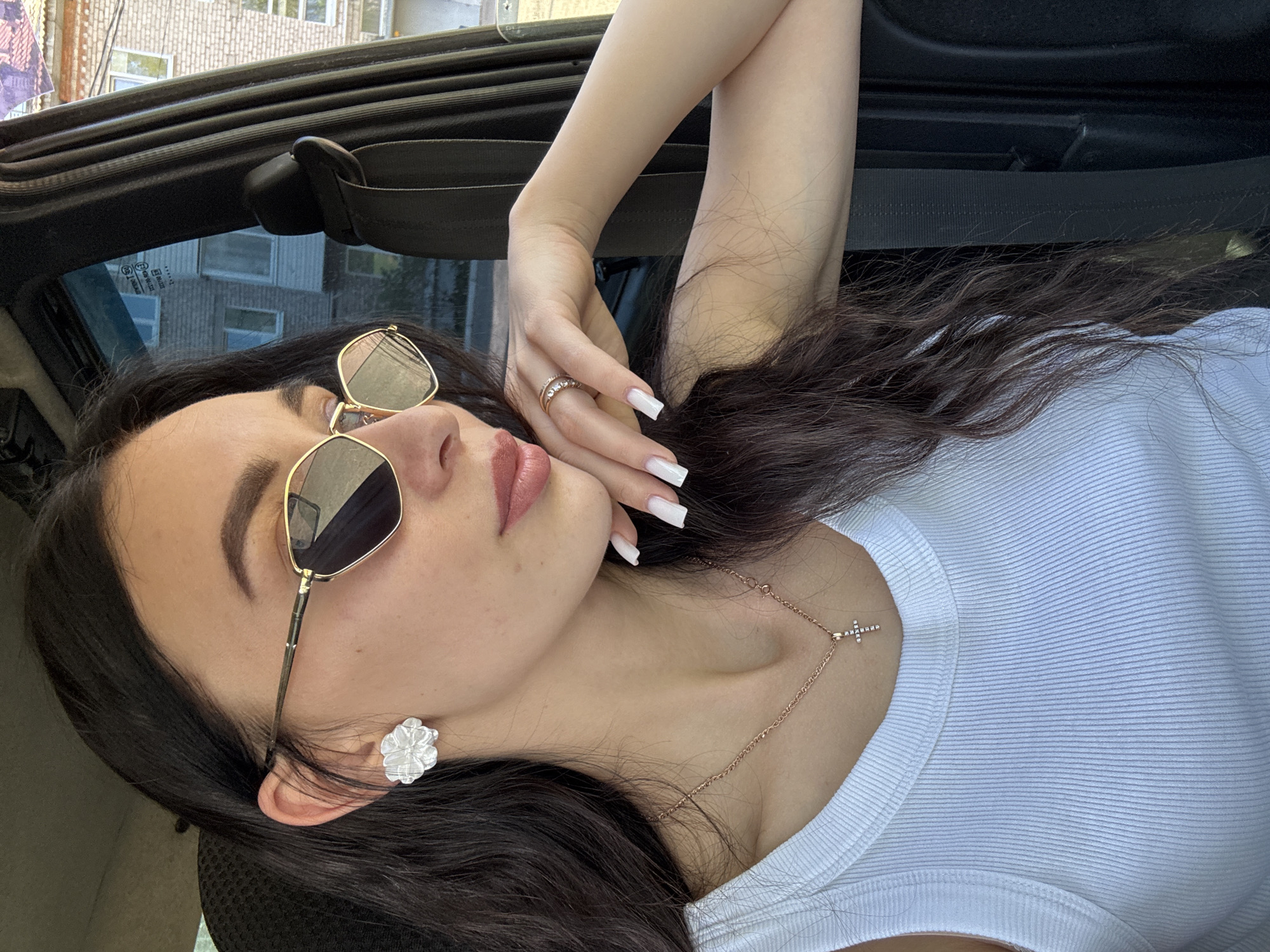 A medium shot depicts a woman lying on her left side, likely within a vehicle, based on the visible car interior. The subject is facing the camera, with dark hair cascading over a white ribbed tank top. She wears large, aviator-style sunglasses with gold frames. A white floral earring adorns her left ear, and a delicate gold necklace with a small cross pendant rests on her chest. Her hands are intertwined near her face, featuring long, square-tipped white nails and a gold ring on her finger. The background reveals a glimpse of buildings through the car window. The location is Chaikovsky, Russia.