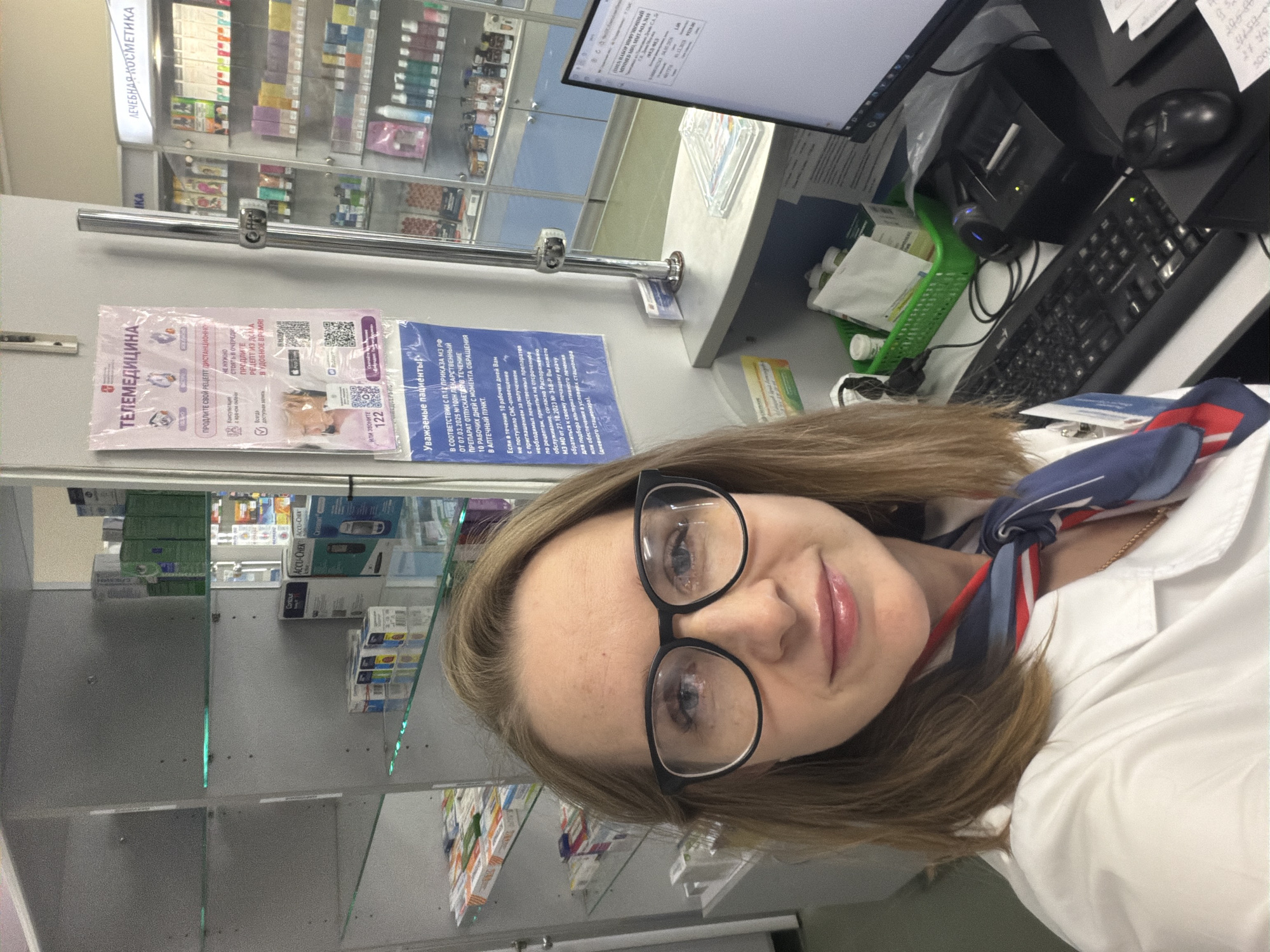 A woman in a white coat and glasses is taking a selfie in what appears to be a pharmacy. Shelves filled with medications and pharmaceutical products are visible behind her. A computer monitor, keyboard, and a printing device are partially visible on a countertop next to her. Posters with information about telemedicine and patient rights are on the wall in the background. The location is possibly in or near Zaprudnya, Russia based on information from the posters in Russian.
