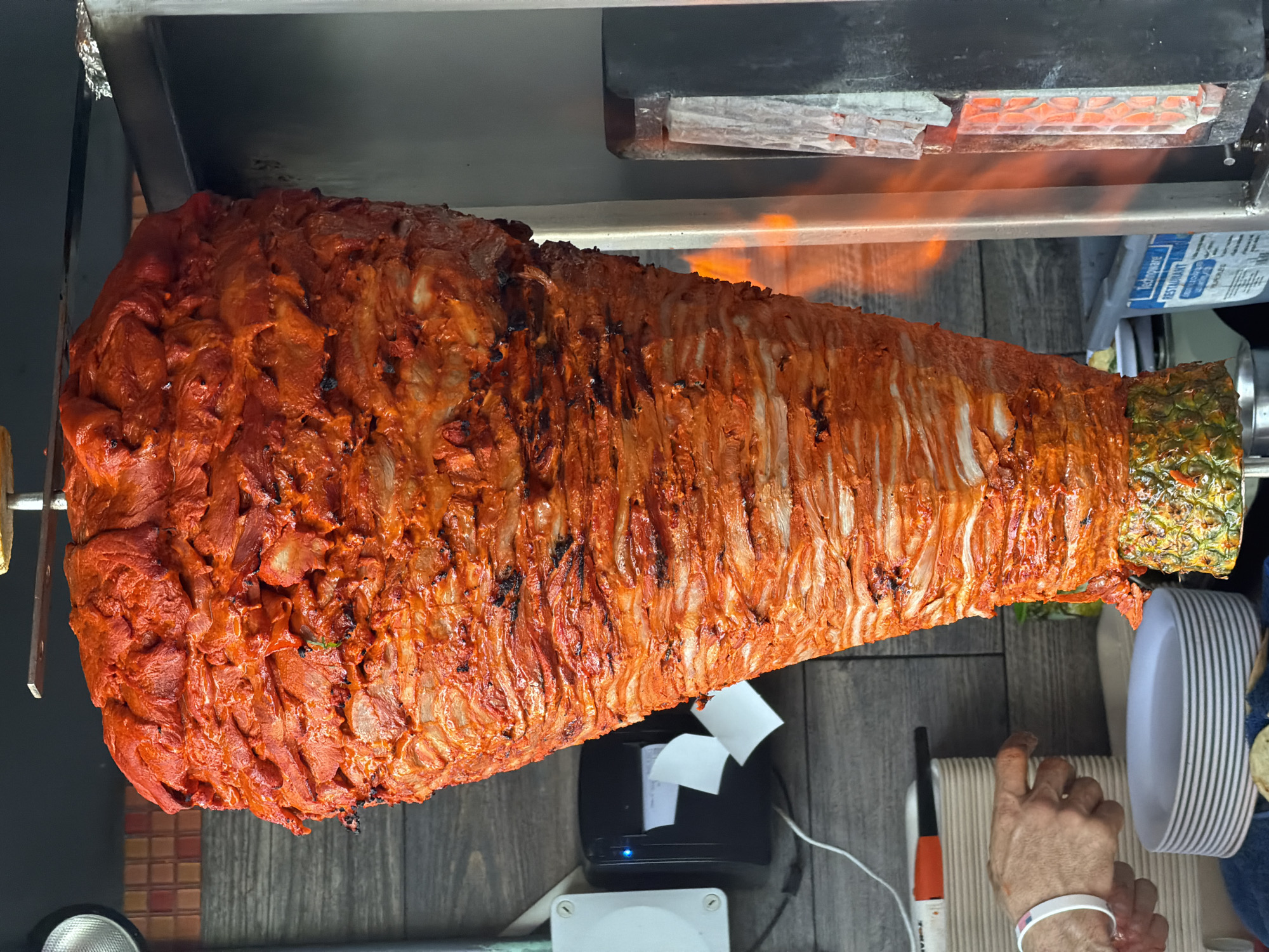 This image depicts a trompo of al pastor meat spinning vertically on a spit, a common sight at taquerias in Cancún, Mexico. The meat, marinated in a red achiote-based sauce, is stacked in thin, overlapping layers, creating a conical shape. The spit is positioned in front of a vertical broiler, evident by the glowing red elements visible to the right, which are roasting the outer layers of the meat, causing some charring and rendering fat. Below the meat, a halved pineapple is impaled on the spit, acting as a base and also flavoring the meat as it cooks.
In the background, a gray, wooden-paneled wall provides a rustic backdrop. To the left, a white electrical box is mounted on the wall, with a black thermal printer attached below it, likely for order tickets. A white power cord runs from the box, and a white roll of paper is partially visible, indicating recent use. A person's hands and forearms are visible in the lower left quadrant of the image. The person is wearing a white wristband and appears to be reaching towards a stack of white plates. A red and black marker pen rests on a stack of white containers behind the plates.
The overall scene suggests an active food preparation environment within a taqueria, likely in Cancún, Mexico, given the prominence of al pastor and the cultural context of such establishments in the region. The lighting appears to be a mix of ambient and artificial light, highlighting the texture and vibrant color of the al pastor. The composition focuses on the trompo as the main subject, with supporting elements of the cooking apparatus and human interaction in the periphery.