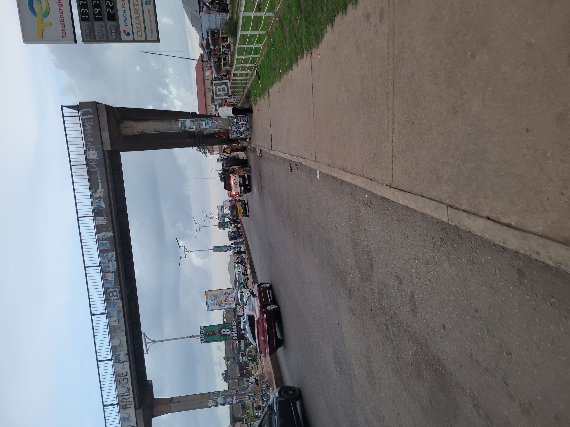 The image depicts a busy urban street scene in an unidentified city. A pedestrian bridge with metal railings and graffiti reading "DR BRIDGE" spans the street, partially obscuring the view of the sky. Along the road, a line of vehicles including cars, trucks, and tricycles are visible, suggesting traffic congestion. A gas station sign indicating prices for petrol, diesel, and kerosene is prominently displayed. On the right side of the road, there's a concrete sidewalk and a grassy patch. Signboards with commercial advertisements are present along the roadside. Pedestrians are seen near the gas station and along the sidewalk.