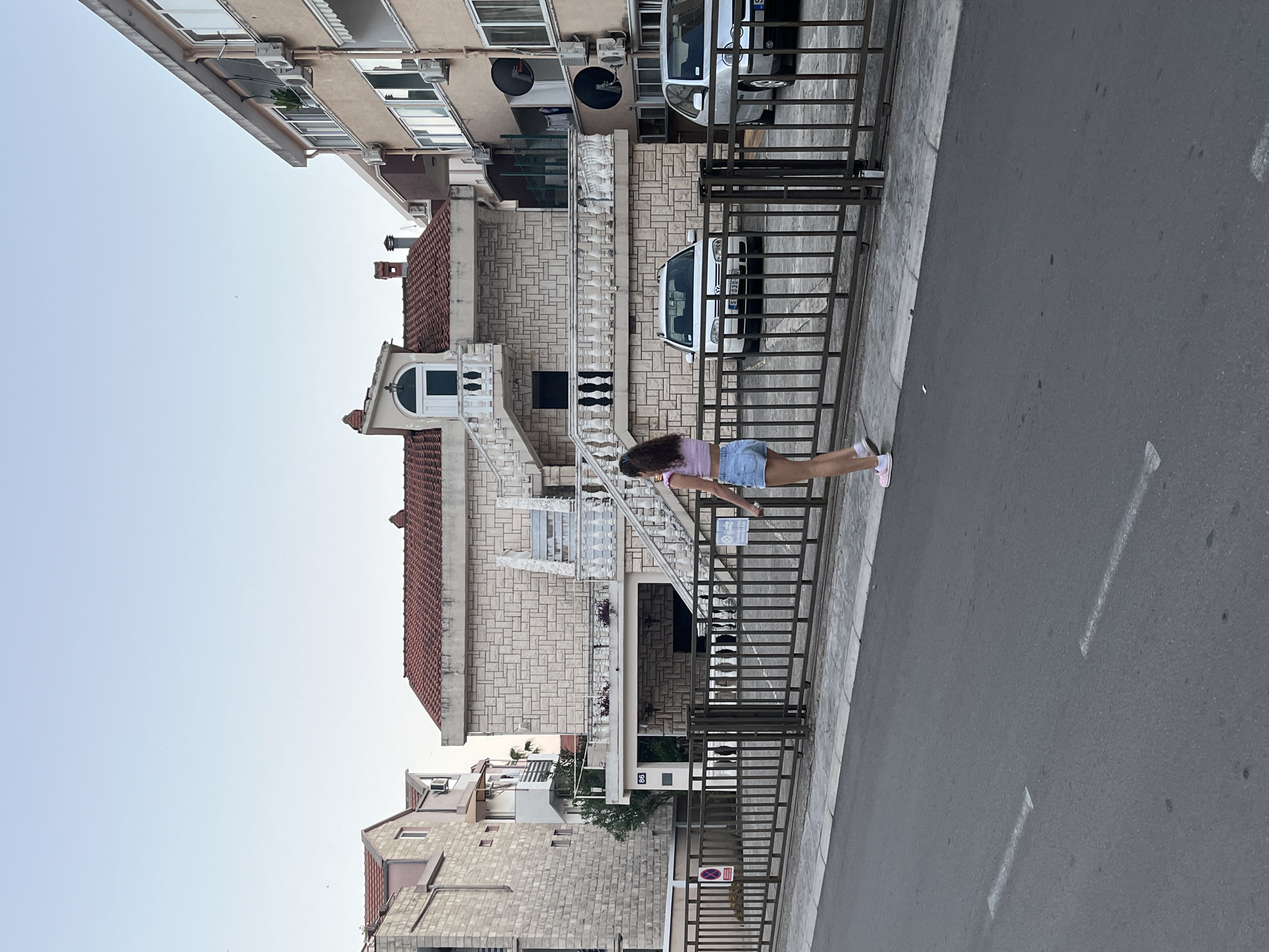 A young woman walks along a sidewalk next to a street in Budva, Montenegro. She is wearing a pink crop top and denim shorts. Behind her, a metal fence separates the sidewalk from a property with a house and two parked cars. The house is multi-story with a light-colored stone facade and a red tile roof. To the left of the house, another building with a similar stone facade is visible. The street in the foreground has white lane markings. The sky is clear and light blue.
