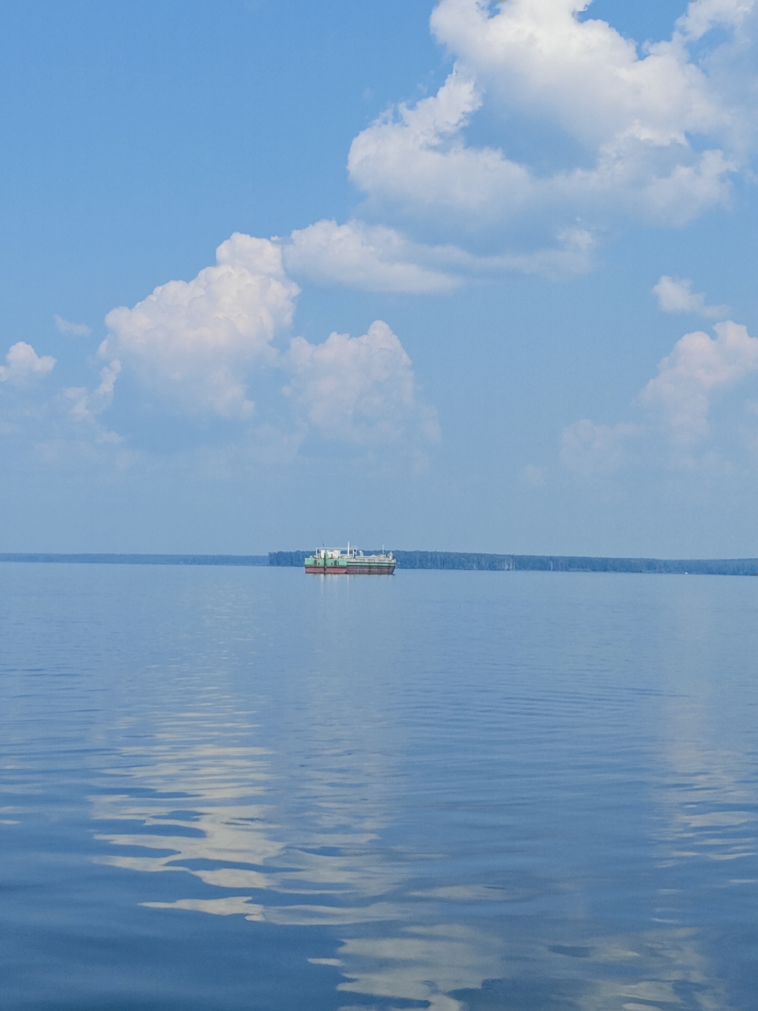 The image shows a wide, calm body of water, likely a reservoir or a large river, reflecting the blue sky and scattered white clouds. In the middle distance, a long, flat-bottomed barge with a white superstructure and a red hull is moving across the water. The barge appears to be a cargo vessel. On the horizon, a dense line of trees suggests a forested shoreline. The scene is tranquil and expansive, with the water dominating the foreground and mid-ground. This setting is consistent with the Kama River or the Votkinskoye Reservoir near the city of Chaikovsky, Russia, known for its wide waterways and surrounding forests. There are no people visible in the image. The overall atmosphere is peaceful and serene, indicative of a quiet day on the water.