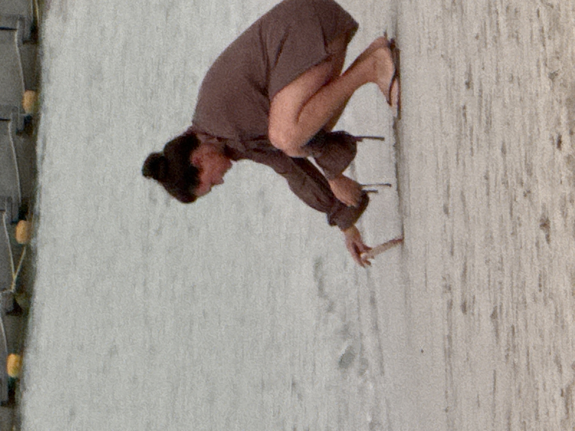 A woman with dark hair pulled into a bun is crouched on a sandy beach. She is wearing a long-sleeved brown robe and flip-flops. Her right hand is extended towards the water, holding a light-colored stick, which she appears to be dipping into the shallow water. Her gaze is directed downwards. The background is a blurry expanse of water.