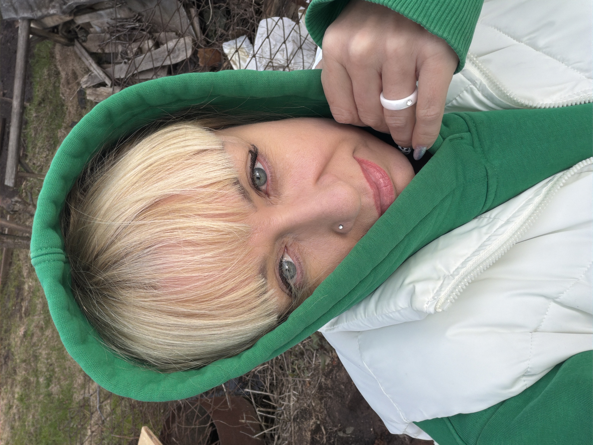 A woman with blonde hair and green eyes is wearing a green hooded sweatshirt and a white puffer jacket. She is looking at the camera with a slight smile. She has a white ring on her finger and a small stud piercing on the right side of her nose. The background shows some outdoor debris including wood and vegetation, possibly indicating a rural area or a less maintained section of a city. The location is Chaikovsky, Russia. The image shows the woman outdoors with debris behind her.