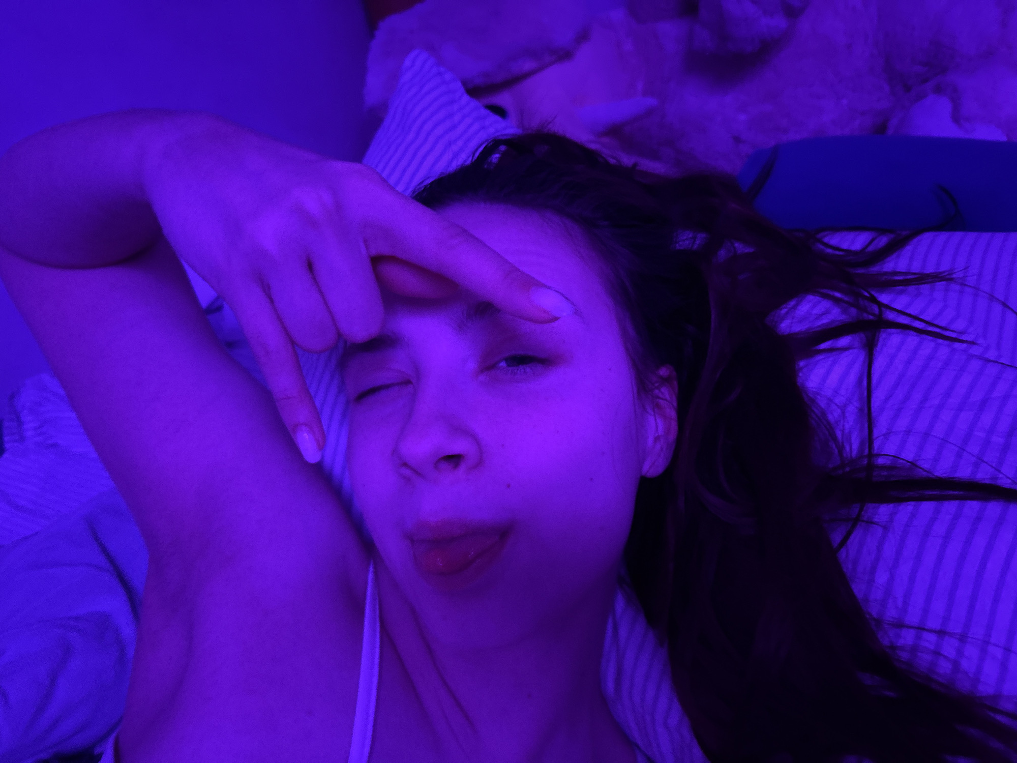 The image depicts a woman lying in bed, illuminated by a strong violet light. She is looking directly at the camera with one eye partially closed and her tongue sticking out. Her right hand is raised, with her index and middle fingers extended and pointed towards her forehead, while her thumb and pinky finger are extended. She is wearing a white tank top. The bed has striped sheets, and there appears to be a stuffed animal in the background. The location in the city of Прикамский, Russia is not discernible from the image.
