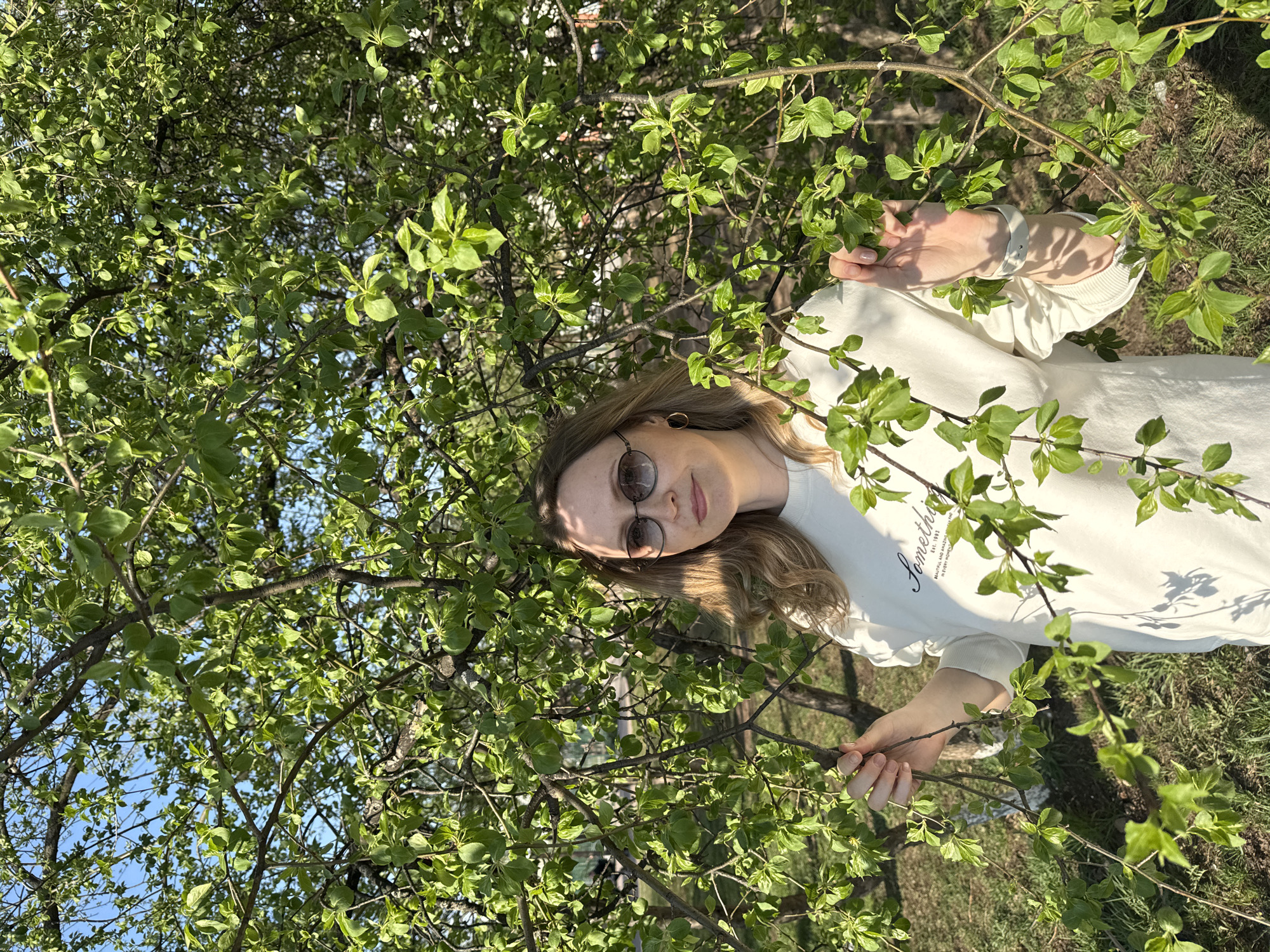 A woman with long blonde hair and round sunglasses stands among branches with green leaves. She wears a light-colored sweatshirt. The photograph appears to be taken outdoors during the day. A patch of grass is visible in the background. The precise location in Chaikovsky, Russia, cannot be determined from the provided image.