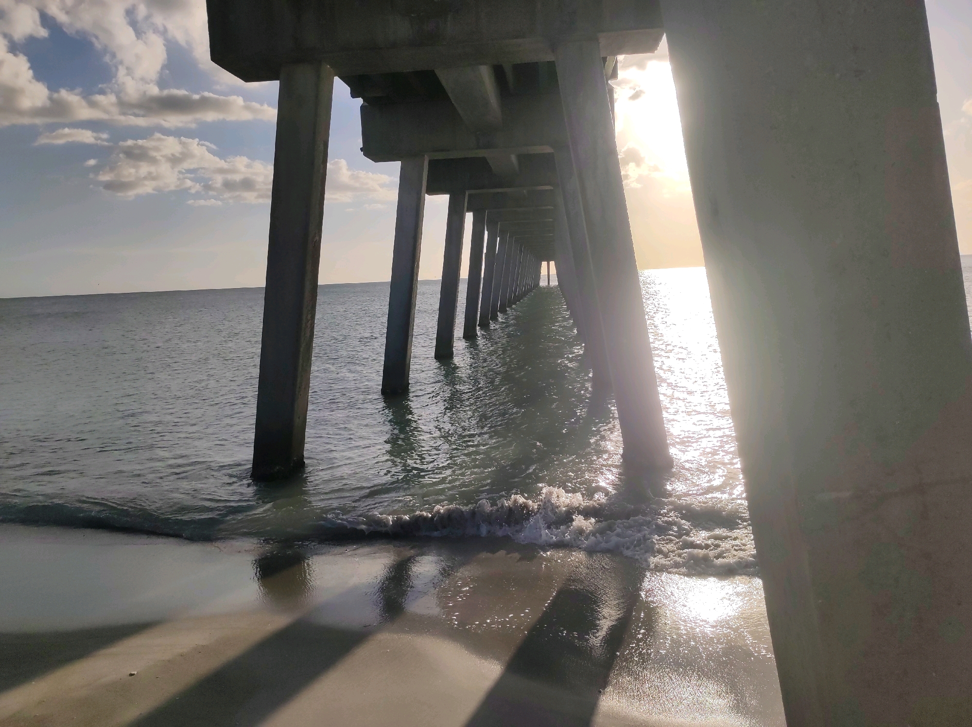 The image depicts a perspective looking out from under a concrete pier towards the ocean during daytime. The pier's supporting pillars, made of concrete, are prominent in the foreground, casting long shadows on the wet sand. Gentle waves are breaking on the shore, with sunlight glinting off the water's surface. The ocean stretches to the horizon, where it meets a pale sky with scattered clouds. The lighting suggests it is either early morning or late afternoon. The scene is located on a beach in Venice, Florida, United States of America.