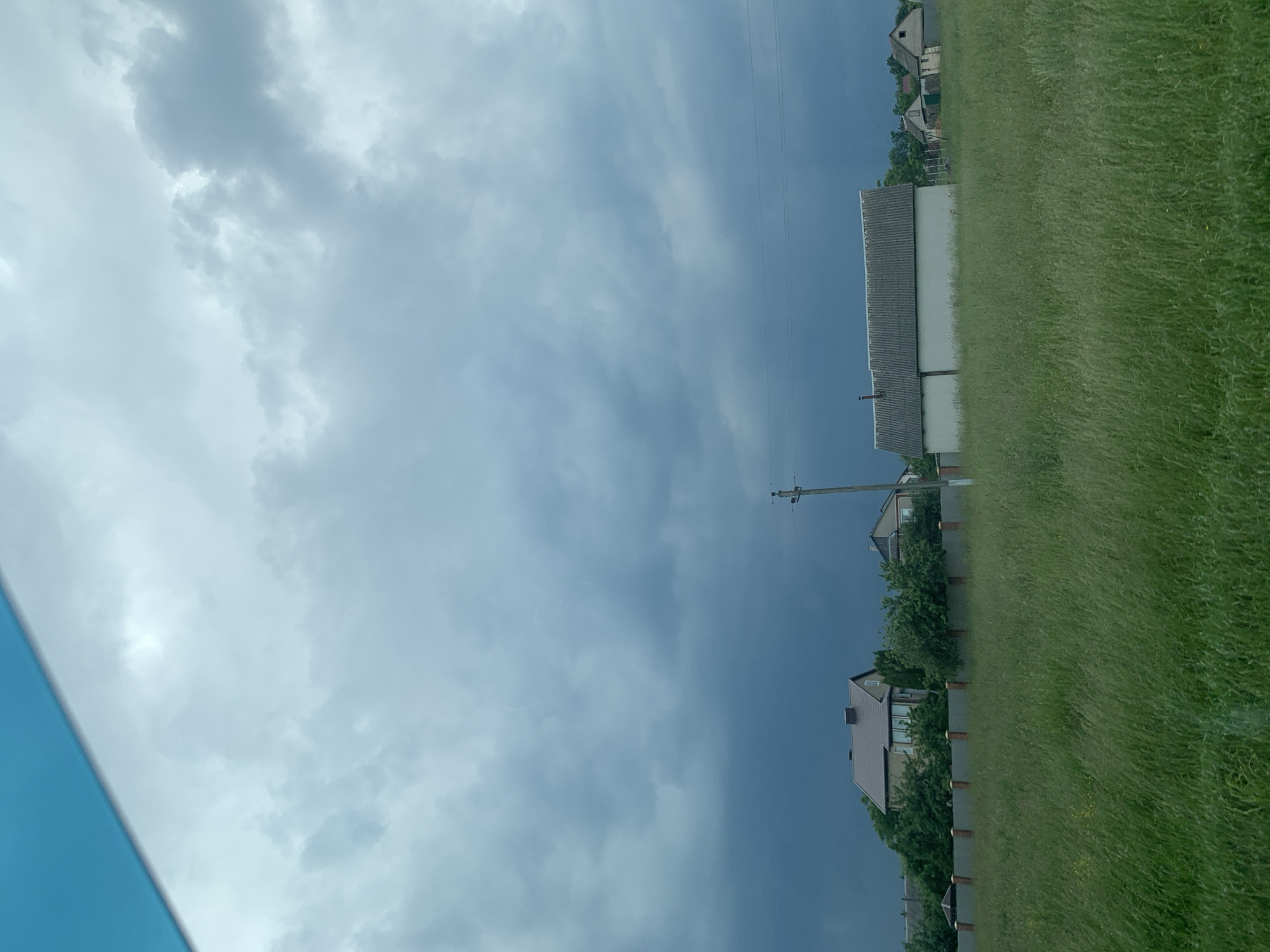 A wide, horizontal shot captures a rural landscape under a dramatic, cloudy sky. In the foreground, tall, green grass sways gently, characteristic of a field. A low, gray fence with intermittent brick posts runs across the mid-ground, separating the field from a row of houses and buildings.
To the left, a two-story house with a brown roof and cream-colored walls is partially obscured by lush green trees. To its right, a long, low building with a gray, corrugated roof and white walls stands prominently. Further to the right, a few smaller houses with pitched roofs are visible, nestled among trees and greenery. A lone utility pole with electrical wires stretches across the scene, adding a touch of man-made infrastructure to the natural environment. The sky above is a mix of dark, stormy gray clouds and lighter, wispy formations, suggesting an approaching storm or the aftermath of one. The overall impression is one of a peaceful, yet potentially unsettled, rural setting. The image is located in Слабада-Кучынскі сельскі Савет, Belarus.