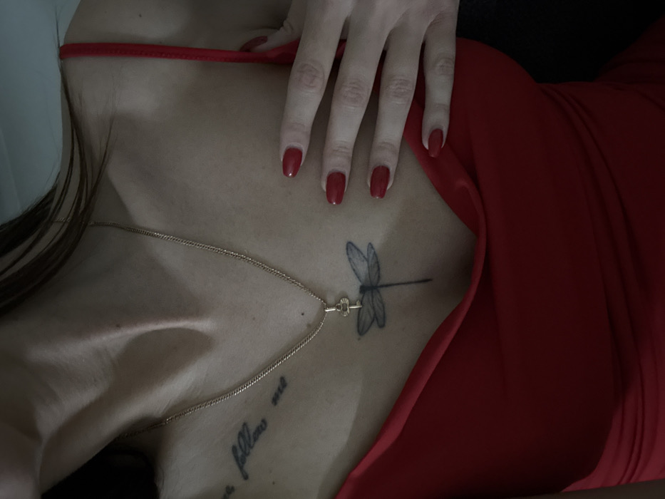 A woman lies angled in a red dress. A delicate gold necklace rests against her skin. She has a dragonfly tattoo and script tattoo on her upper chest. Her hand, with red nail polish, rests on her upper body. The angle is shot from above, close-up. The light is soft. The background is dark, emphasizing the subject.