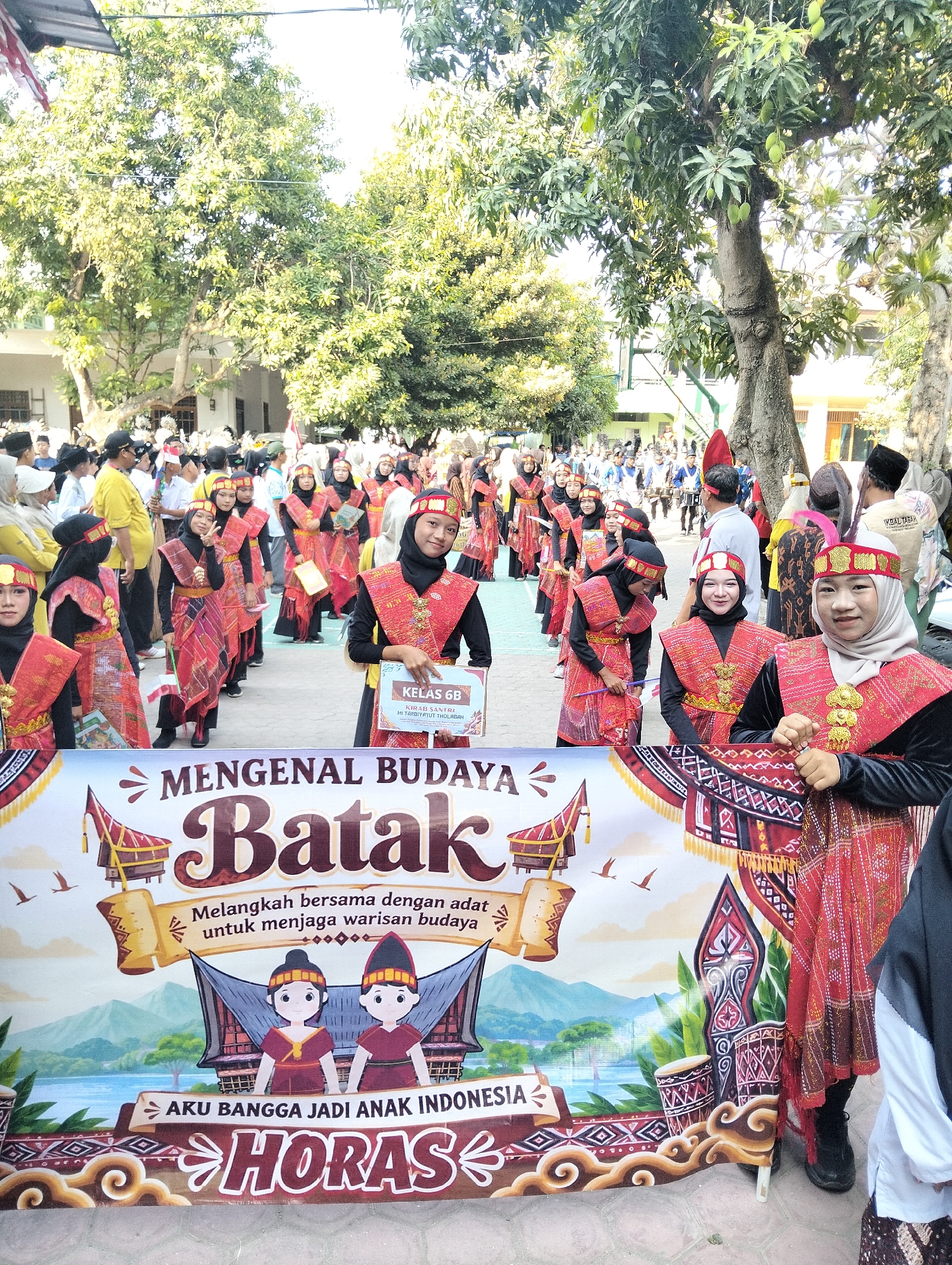 In Kranji, Indonesia, a group of young people, predominantly female, are participating in a cultural parade or event. They are dressed in traditional Batak attire, characterized by vibrant red and black patterned fabrics, gold embellishments, and head coverings. Many are wearing matching outfits and headbands. Some individuals hold small Indonesian flags or awards. In the foreground, a large banner displays the text "MENGENAL BUDAYA Batak" (Introducing Batak Culture) and other Indonesian phrases, along with colorful illustrations of traditional Batak houses, musical instruments, and symbolic motifs. The backdrop suggests an outdoor setting, likely a school or public space, with trees and buildings visible. The overall atmosphere appears festive and celebratory, showcasing cultural pride.