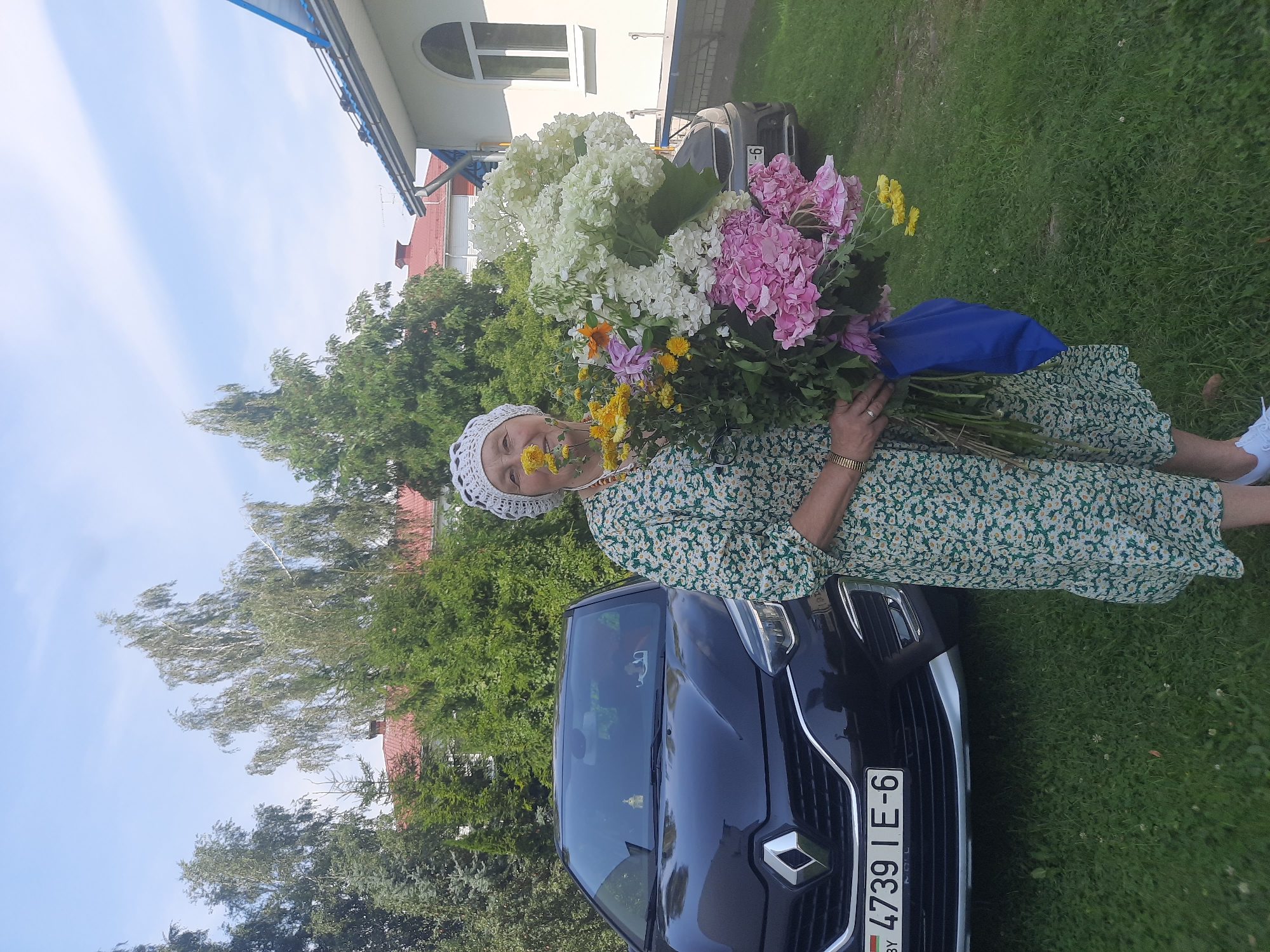 An elderly woman wearing a floral green dress and a white crocheted hat stands outdoors holding a large bouquet of flowers. She is positioned in front of a black Renault car with a license plate that reads "4739 IE-6 BY." The background includes green trees, a building with a blue-tiled roof, and a clear blue sky with some clouds. The woman appears to be sniffing a yellow flower from the bouquet. The scene is set on a grassy area, suggesting a residential or park-like setting. The image was taken in the city of Shklov, Belarus.