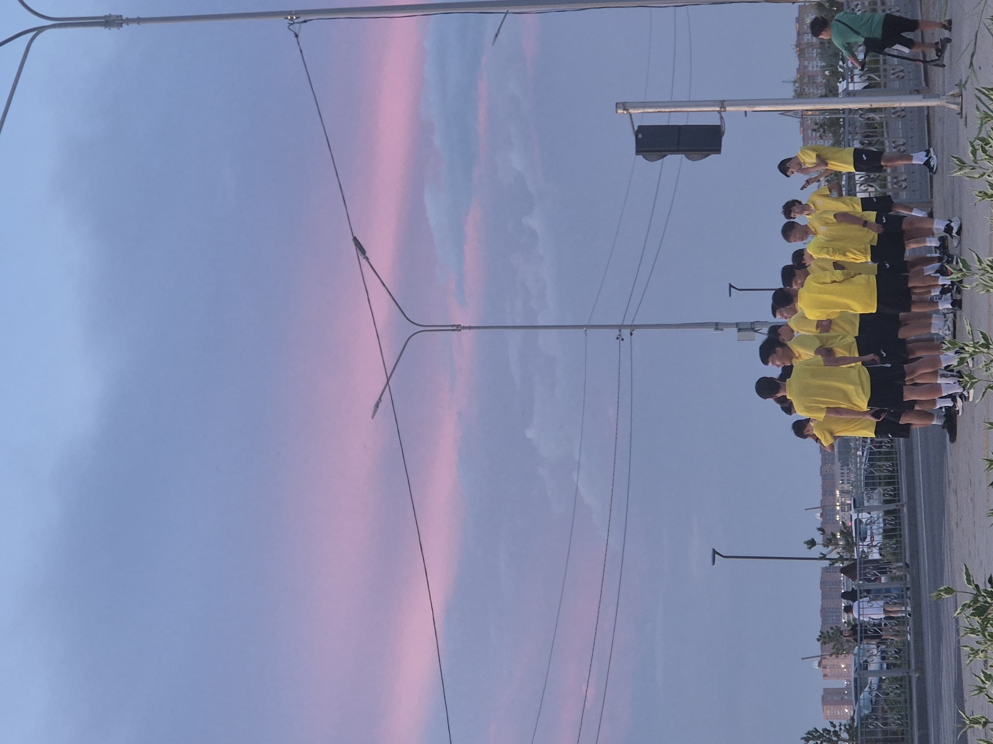 A group of young males, appearing to be teenagers, are gathered on a sidewalk next to a street. Most of them are wearing bright yellow t-shirts and black shorts. One individual in the foreground on the right is wearing a green shirt and riding a scooter. In the background, there is a cityscape with buildings, a parking lot with cars, and a bridge or overpass. Overhead, there are streetlights and power lines against a sky with pink and blue clouds, indicative of either sunrise or sunset. The scene appears to be in an urban environment in Kokshetau, Kazakhstan, likely near a main road or intersection, given the presence of traffic lights and street infrastructure.