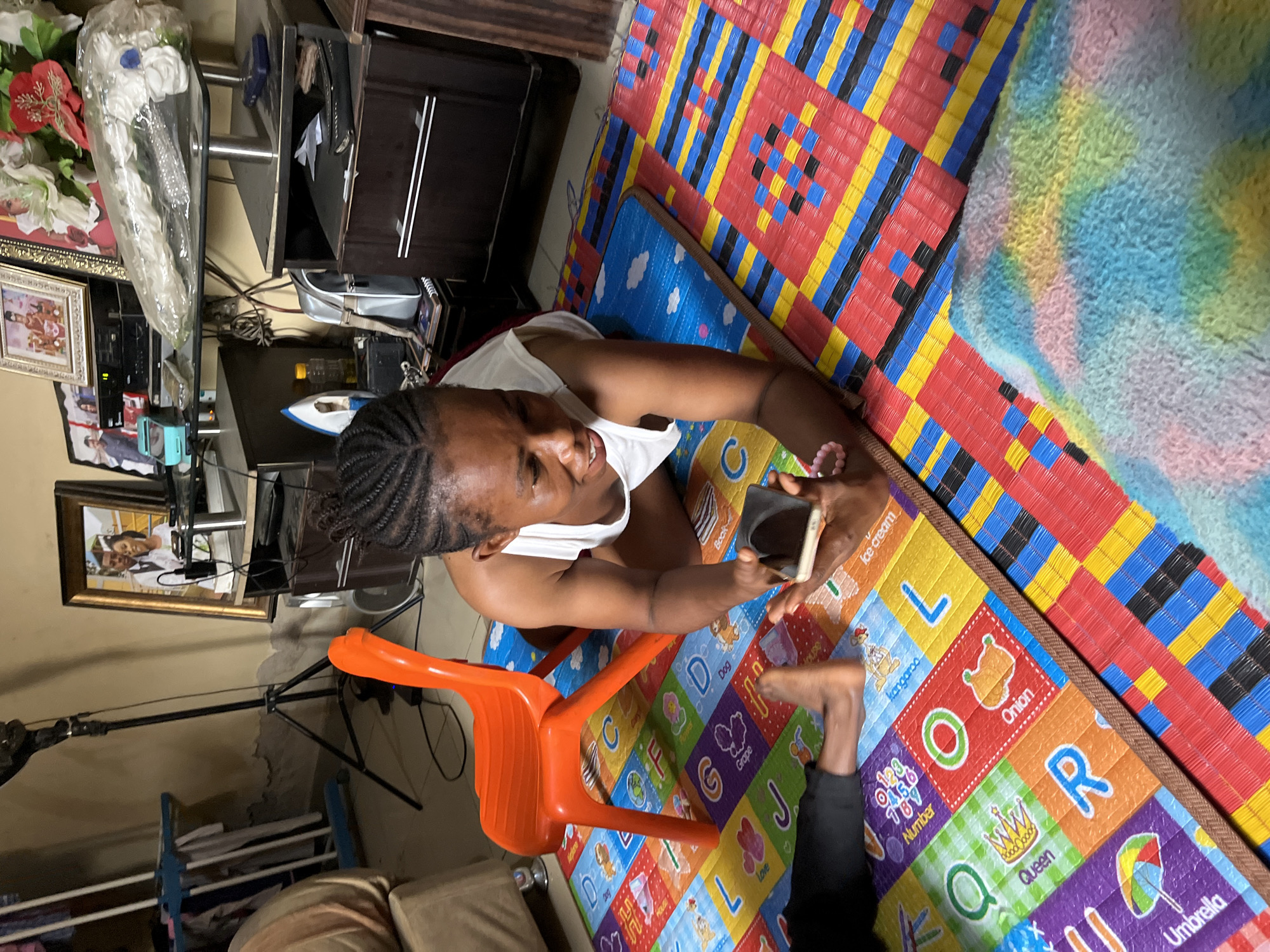 A woman is seated on an orange chair while holding a smartphone, seemingly looking at its screen. The chair is placed on a colorful alphabet play mat, which is laid on top of a more vibrant, multi-colored mat. A portion of another person's leg is visible on the play mat. Beside the play mat is a fuzzy, multi-colored rug.
In the background, there is a multi-tiered shelf unit containing various items including an iron, framed photos, and decorative flowers in a plastic bag. A camera tripod stands to the left. This scene is located in a room in Madalla, Nigeria.