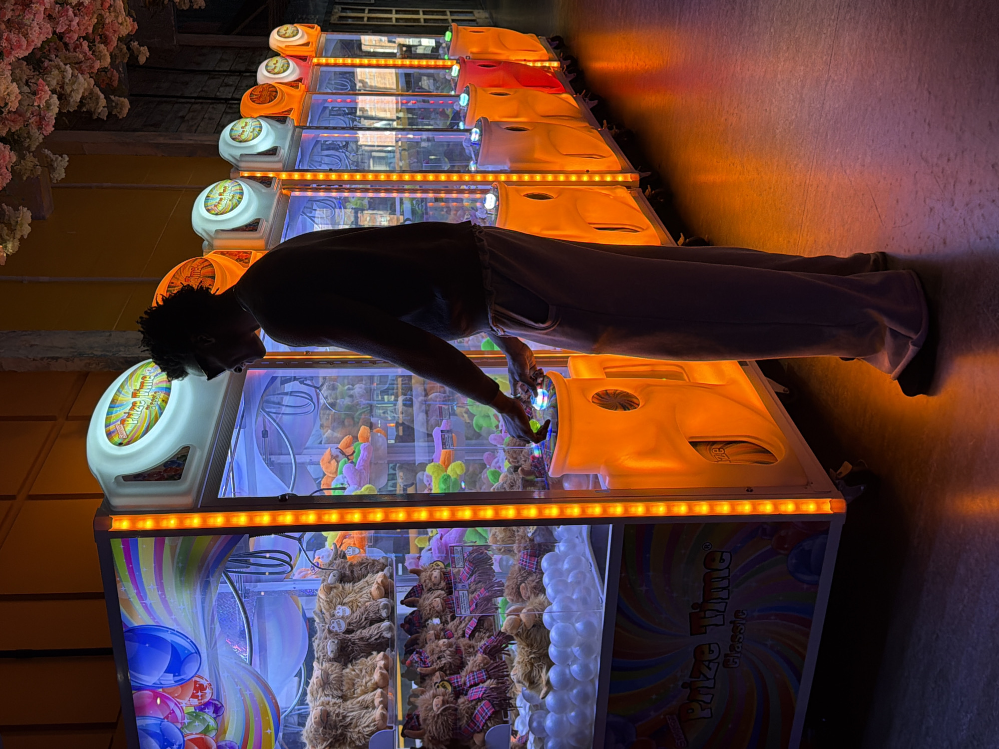 A person in a dark top and light trousers leans over a row of "Prize Time Classic" arcade claw machines. The machines are illuminated with colorful lights and filled with plush toys. The location is unknown, but it is not in London, United Kingdom.