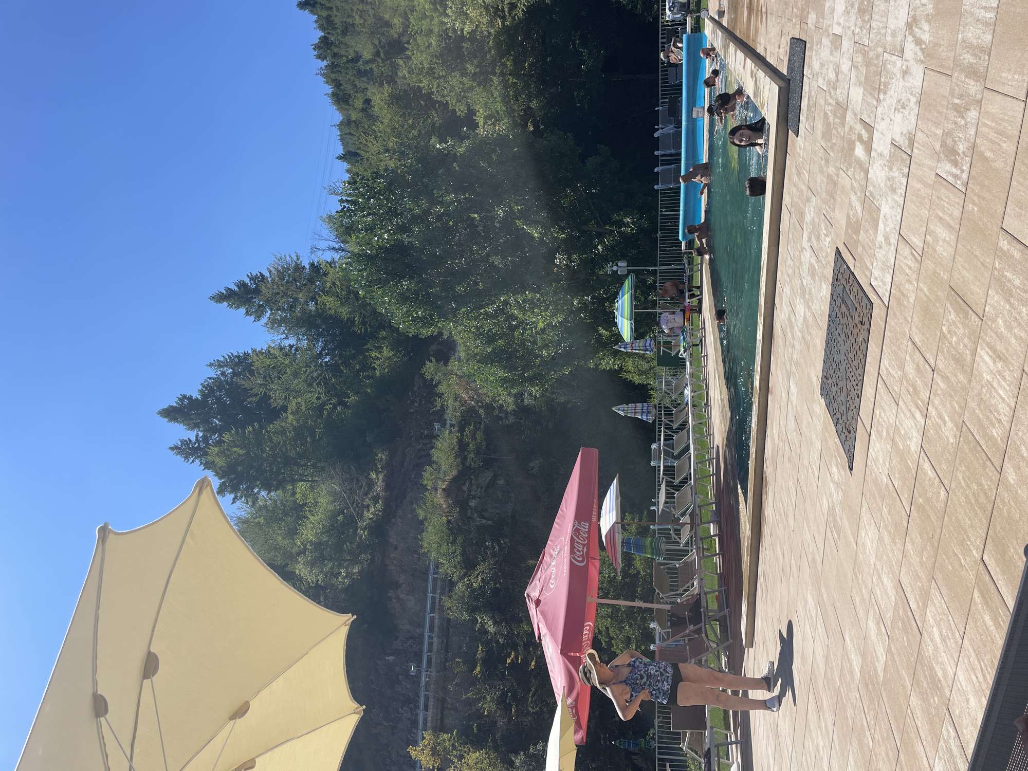 The image depicts an outdoor swimming pool area on a sunny day, located in Banite, Bulgaria. In the foreground, a woman wearing a patterned swimsuit and a wide-brimmed hat stands on a tiled patio, adjusting her attire. To her left, a large, light-colored umbrella shades a portion of the patio. Behind her, a bright pink umbrella with "Coca-Cola" branding is visible, accompanied by several lounge chairs.
The main subject of the image is the rectangular swimming pool, filled with clear blue water. Several people are in the pool, including adults and children, some of whom are swimming and interacting. The pool is surrounded by a white fence, and more lounge chairs are arranged on the patio.
The background consists of a steep, tree-covered hillside with a road running along it. The trees are lush and green, indicating a natural, possibly mountainous, environment. The sky is a clear, bright blue. The overall scene conveys a relaxed, recreational atmosphere typical of a summer resort or public swimming facility.