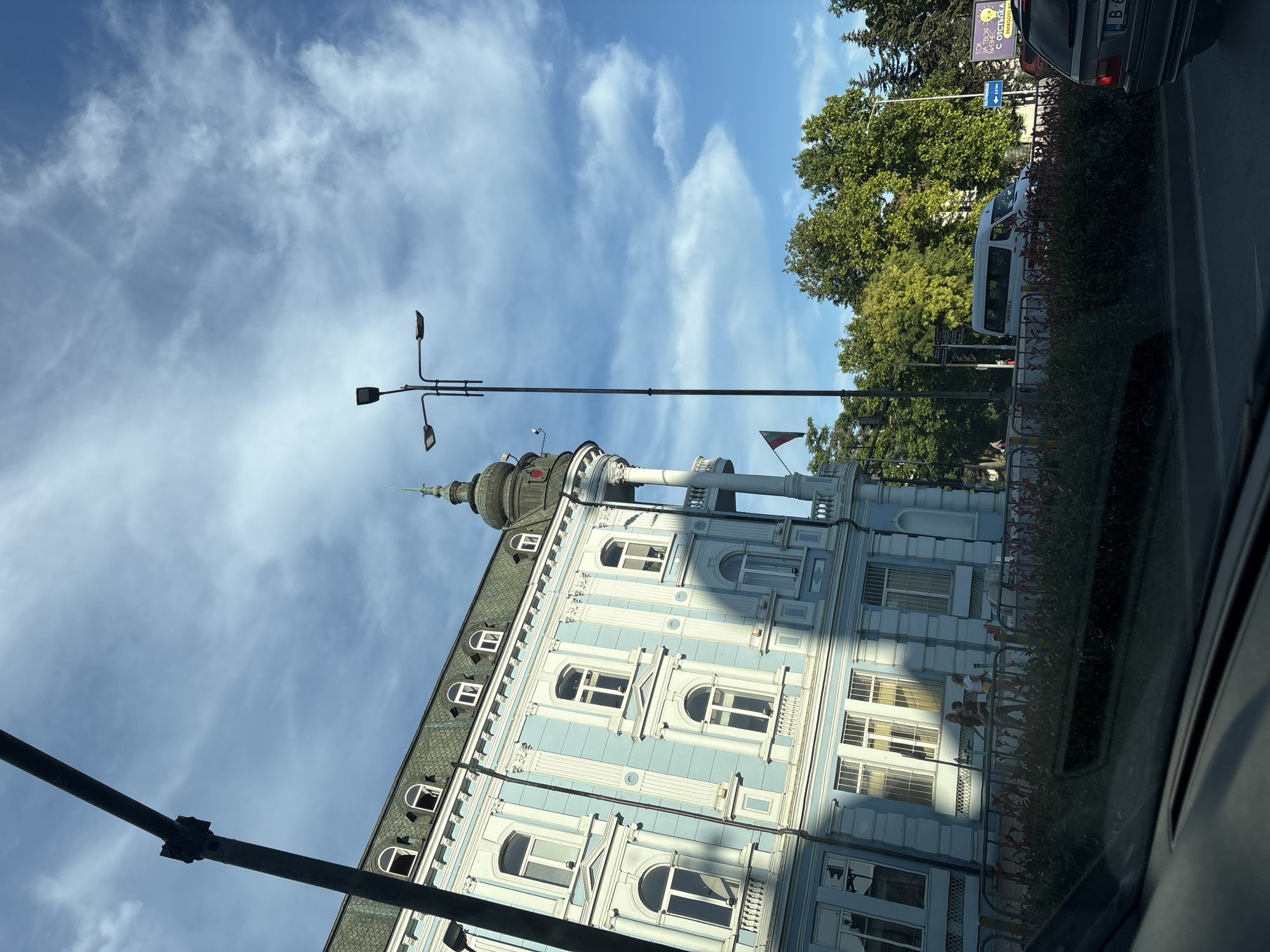 The image captures a street view in Varna, Bulgaria, featuring a light blue building with white trim and ornate architectural details. The building has a mansard roof with dormer windows. A streetlamp with multiple lights stands prominently in the foreground, partially obscuring the sky, which is blue with scattered white clouds. Green trees line the background. In the middle ground, people are walking along a sidewalk behind a decorative fence with flowering plants. A white van is visible on the street, and a dark-colored car is in the far right of the frame. A flag, partially visible, hangs from the building. The scene appears to be a public area within the city.