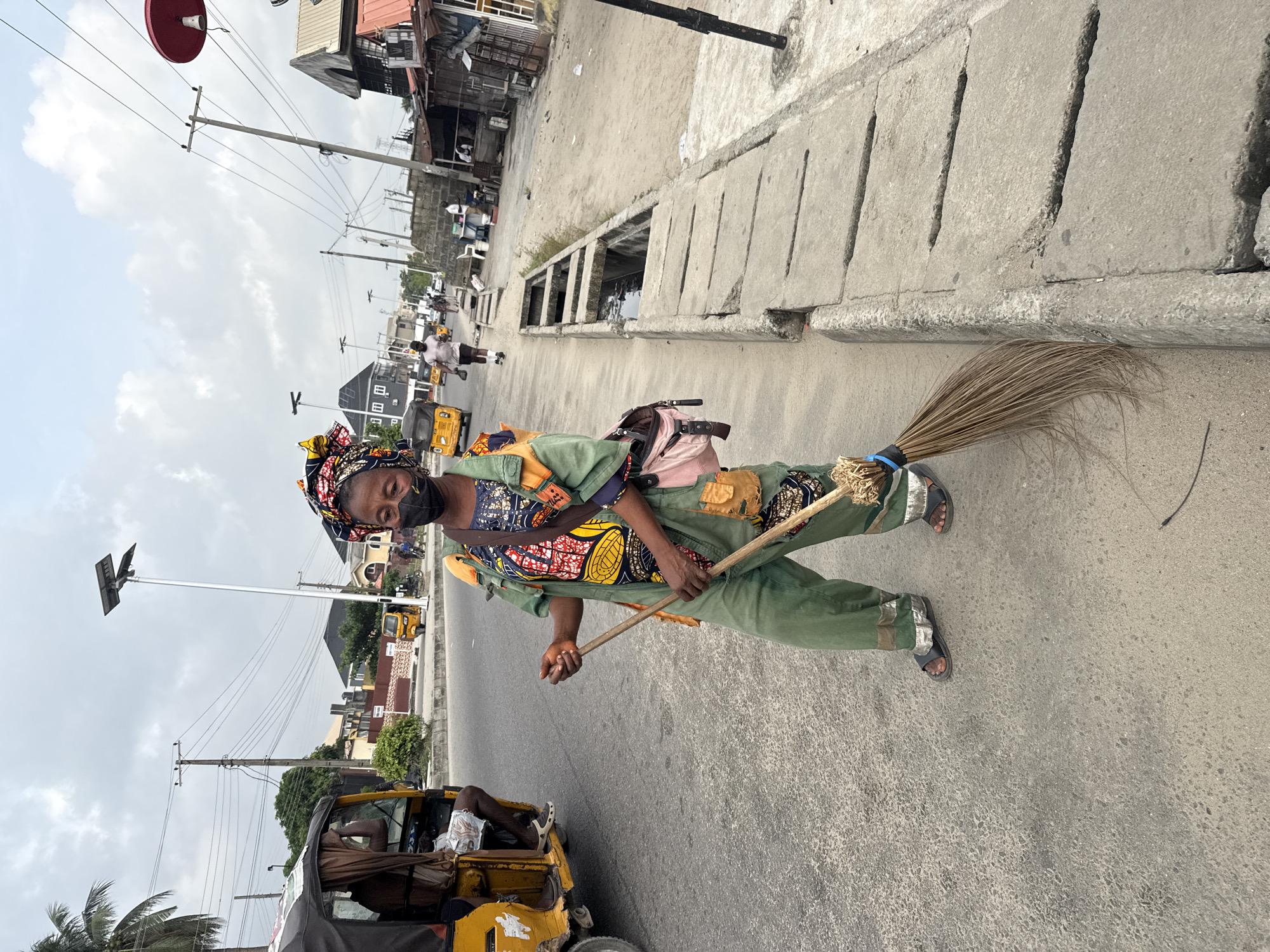 A woman wearing a green uniform, colorful headwrap and a face mask is standing on the side of a road in Ado, Nigeria, holding a broom. She is dressed in an outfit that includes patches of patterned fabric. The broom has a natural fiber head bound with a blue tie. Behind her is a yellow three-wheeled vehicle, or 'keke,' with a partially visible passenger. The road is grey, and a concrete curb and drainage system are visible to her right. In the background, there are buildings, overhead power lines, street lights, and pedestrians in the distance. The sky is overcast.