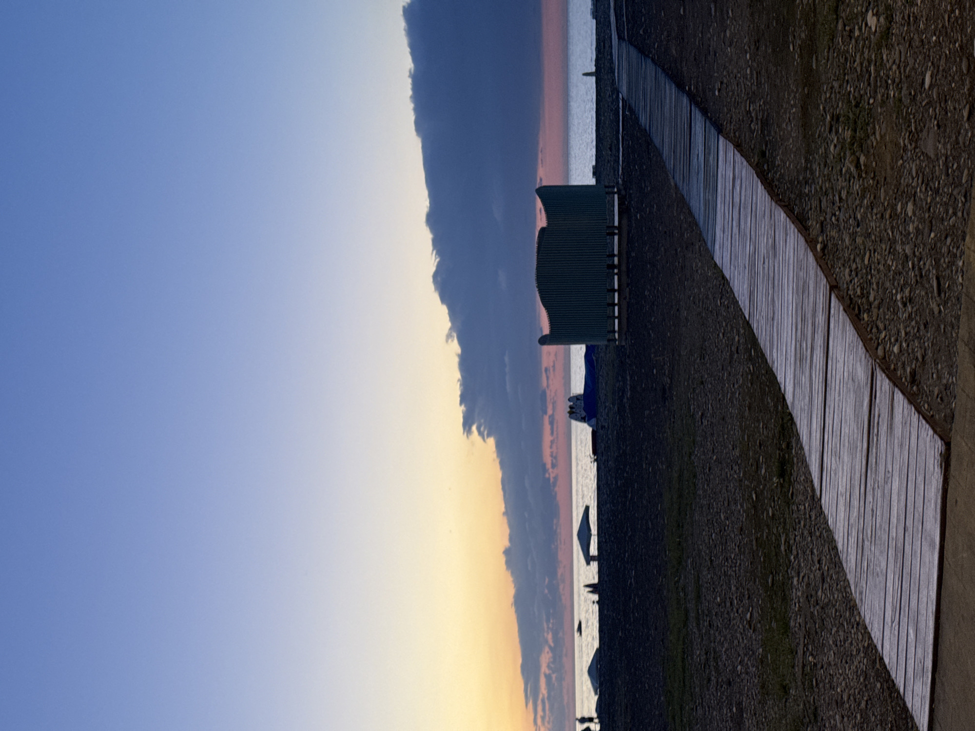 The image depicts a beach scene at dusk, likely on the Batumi coast in Georgia. The sky transitions from a pale blue at the top to soft oranges and pinks near the horizon, indicative of sunset. A large, dark cloud formation hovers over the sea, partially obscuring the colorful sky.
In the foreground, a wooden boardwalk curves from the bottom right of the frame towards the sea, laid across a pebble-strewn beach. To the left of the boardwalk, a dark, corrugated structure, possibly a changing room or restroom, stands on stilts. Further back, near the water's edge, several white beach umbrellas are visible, some casting shadows. A few figures, silhouetted against the sea, are walking on the beach. On the water, a small boat is seen in the distance. The overall atmosphere is calm and serene, with the fading light creating a tranquil ambiance.