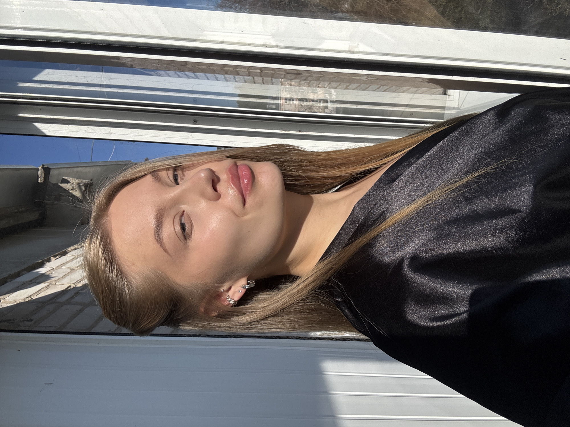 A fair-skinned young woman with blonde hair is positioned indoors, possibly near a window. Her head is tilted back, and she is wearing a black garment. Her face is illuminated by sunlight, highlighting her features. It's difficult to definitively identify the location as Chaikovsky, Russia based solely on this image. The interior setting appears to be simple, and the view from the window shows an exterior with undefined elements.