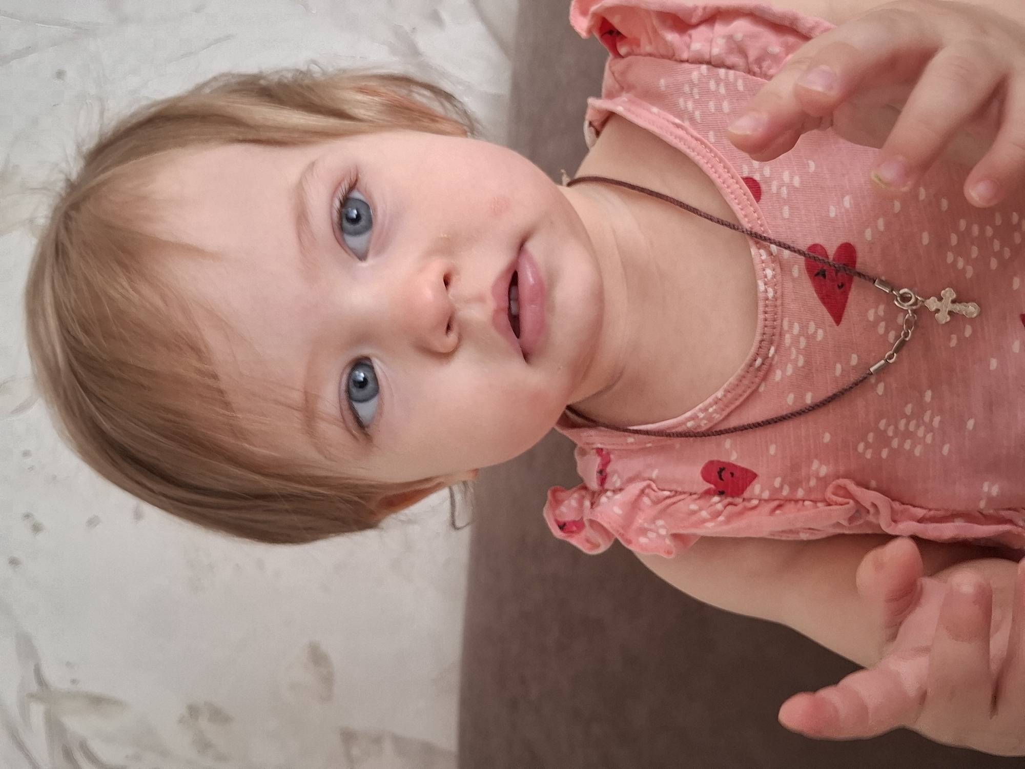 The image is a portrait of a baby or toddler, captured with a slightly rotated perspective. The child has fair skin, bright blue eyes, and light brown hair. They are looking directly at the camera with an open mouth, conveying a sense of curiosity and engagement. A small, reddish mark is visible on their left cheek.
The baby is wearing a pink top patterned with numerous small white polka dots and several larger red hearts. Around their neck, a thin brown cord necklace is visible, from which a small, silver-colored cross pendant hangs. The baby's hands are also visible; one is raised near their face with fingers slightly splayed, and the other rests lower near their chest.
The background features a light, textured surface on one side and a darker, plain surface on the other, suggesting an indoor home environment such as a bed or sofa. The lighting is soft and natural, consistent with daytime indoor conditions. No specific location cues for Chaikovsky, Russia, are discernible within the image itself. There is no visible text. The overall impression is one of innocent charm and direct interaction with the viewer.