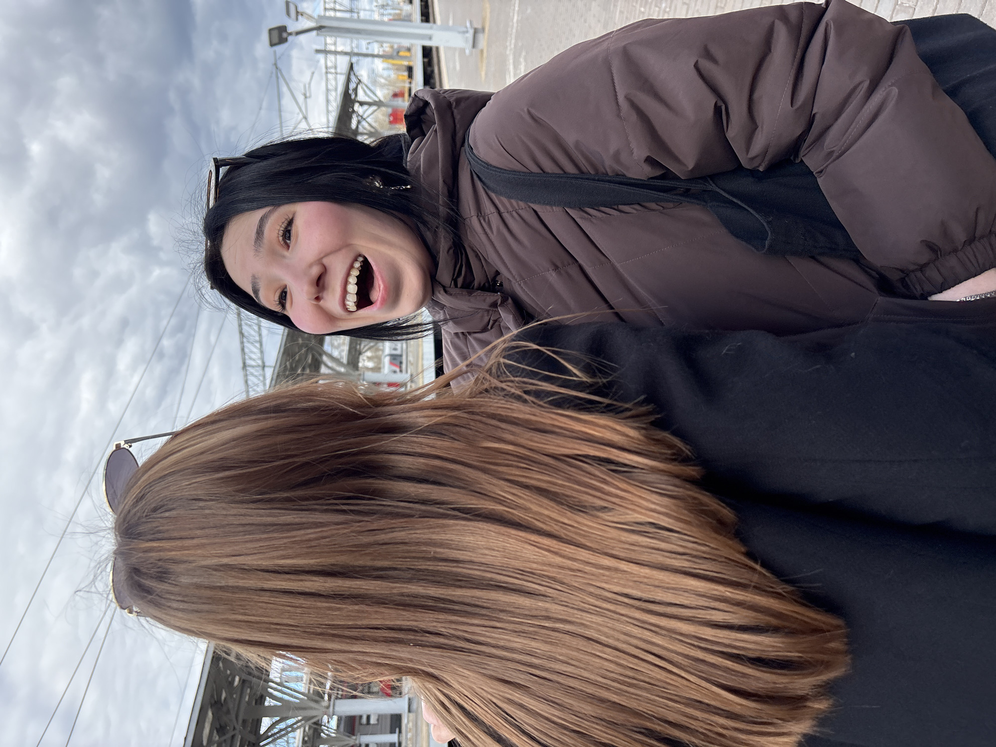 Two women are posing for a photo at what appears to be a train station in Kazan, Russia, based on visible signs and architecture. One woman has long, dark brown hair and is wearing sunglasses; her face is partially obscured. The other woman is fair-skinned with black hair and is wearing a brown puffer jacket. She's laughing with her mouth open, and sunglasses are perched on her head. The background shows station infrastructure, including metal structures, overhead wires, and a cloudy sky.