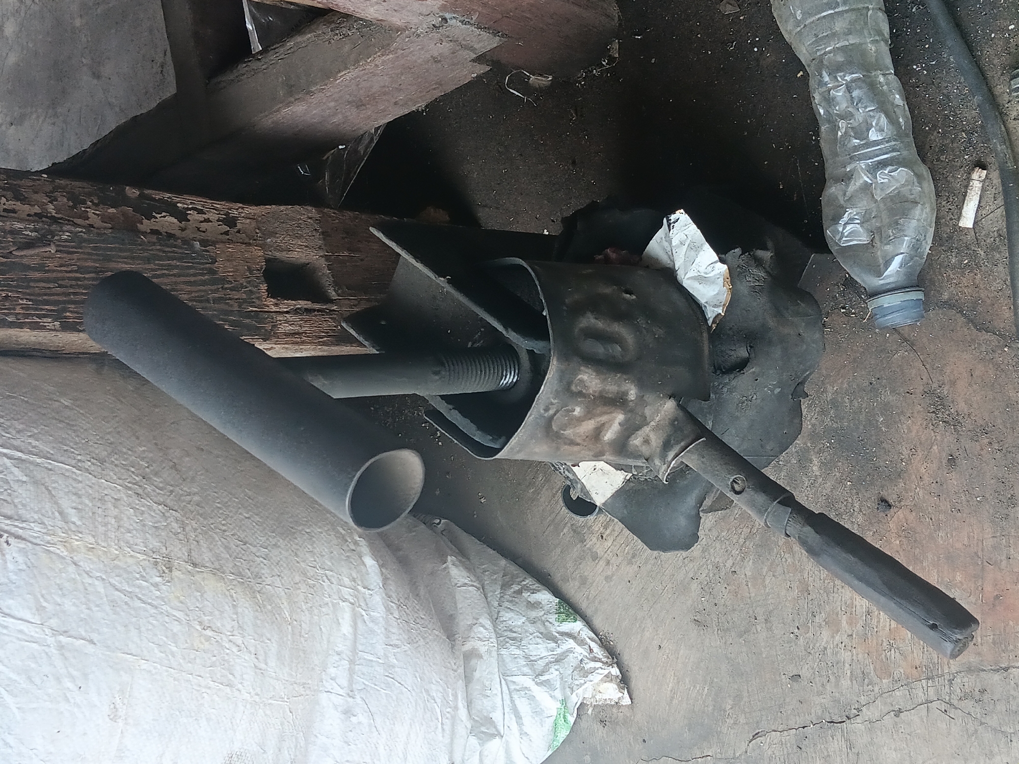 The image shows a close-up, eye-level view of a discarded tire and a metal object, possibly a manual press or clamp, on a concrete floor. To the left, a large, crumpled white sack is visible. Behind the metal object, aged wooden beams form a rough structure, suggesting an outdoor or semi-enclosed workspace. A clear plastic bottle lies on the ground in the foreground, to the right of the metal object, and a cigarette butt is situated just below it. The scene appears to be an outdoor area, likely a workshop or storage space, in the village of Batursari, Indonesia.