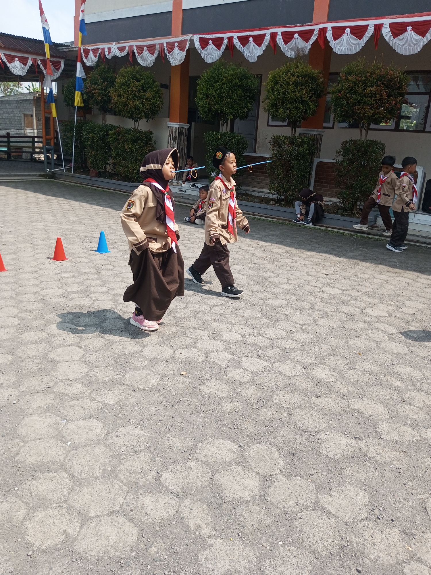 In Kalibeber, Indonesia, children in scout uniforms participate in an outdoor activity. Two children in the foreground, a girl on the left wearing a brown hijab and a brown skirt, and a boy on the right wearing brown pants, walk across a grey hexagonal paved ground. The girl is holding a blue straw in her mouth. Behind them, other children in similar uniforms are visible, some sitting and some standing. In the background, a building with a grey facade and orange trim is adorned with red and white decorative banners. Several green bushes and trees are planted in front of the building. Orange and blue cones are placed on the ground, suggesting a game or obstacle course. Red and white flags are mounted on poles to the left of the building. The scene is illuminated by bright sunlight, casting shadows on the paved ground.
