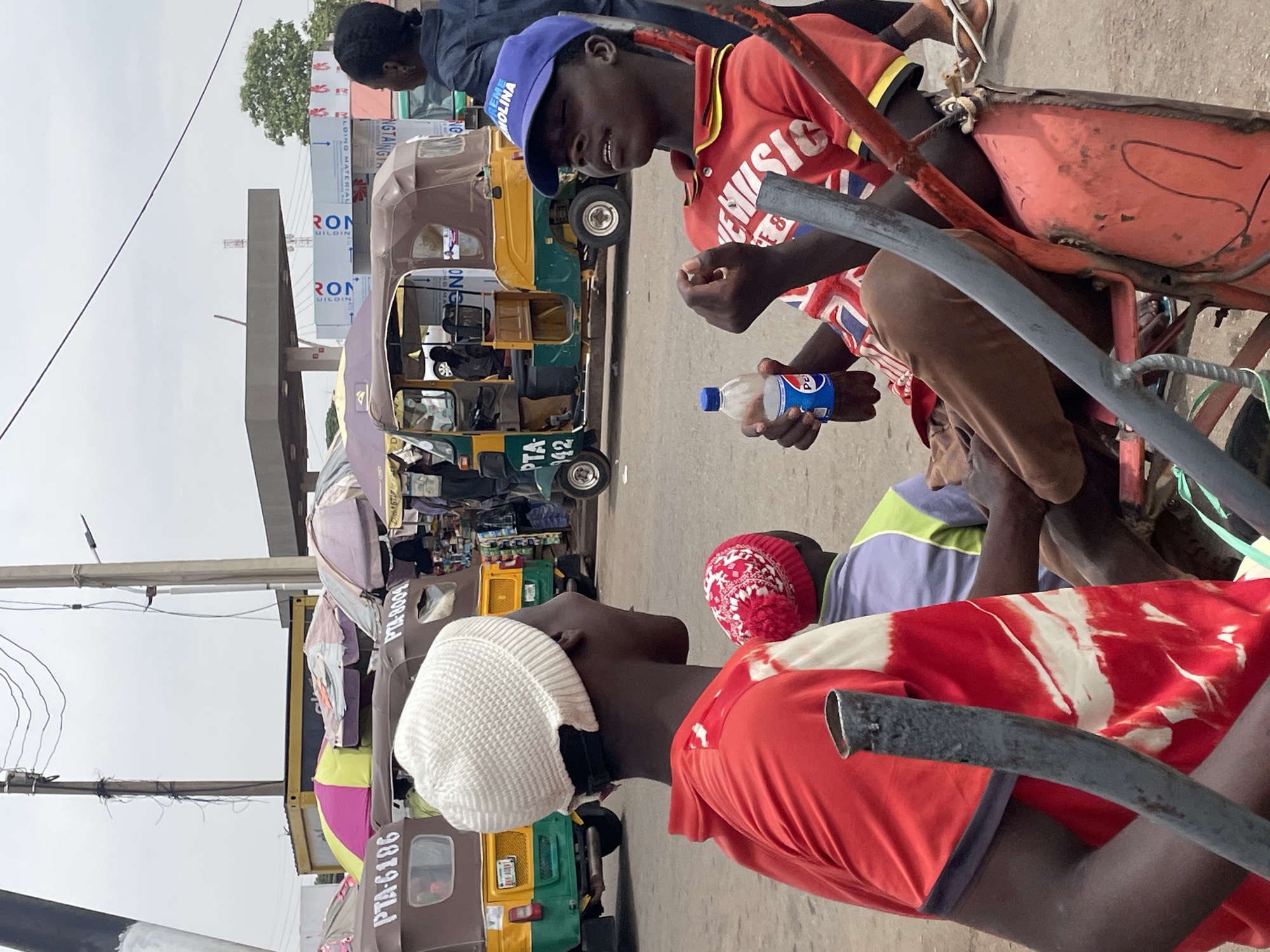 The image captures a street scene in what appears to be Jos, Nigeria, where several auto-rickshaws (tuk-tuks) are parked. Three young men are visible in the foreground. One is seated on what appears to be a hand-pulled cart. He is wearing a red shirt with the word "EMUSIC" printed on it, and a blue baseball cap with the word "MOLINA" printed on it. He is holding a small, blue and white "Pepsi" bottle. A second young man, wearing a red hat, is sitting on the ground next to him. The third young man in a white hat is standing.
Behind them are several tuk-tuks with license plates partially visible, including "PTA-6186," "PTA-8009," and "PTA-142." Buildings and signage in the background suggest commercial establishments, including a sign that reads "RON UILDIN" and "MLDING MATERIAL". The setting appears to be an urban street or roadside area. The scene conveys a casual moment of rest or interaction amongst the young men amidst the everyday backdrop of transportation and commerce.