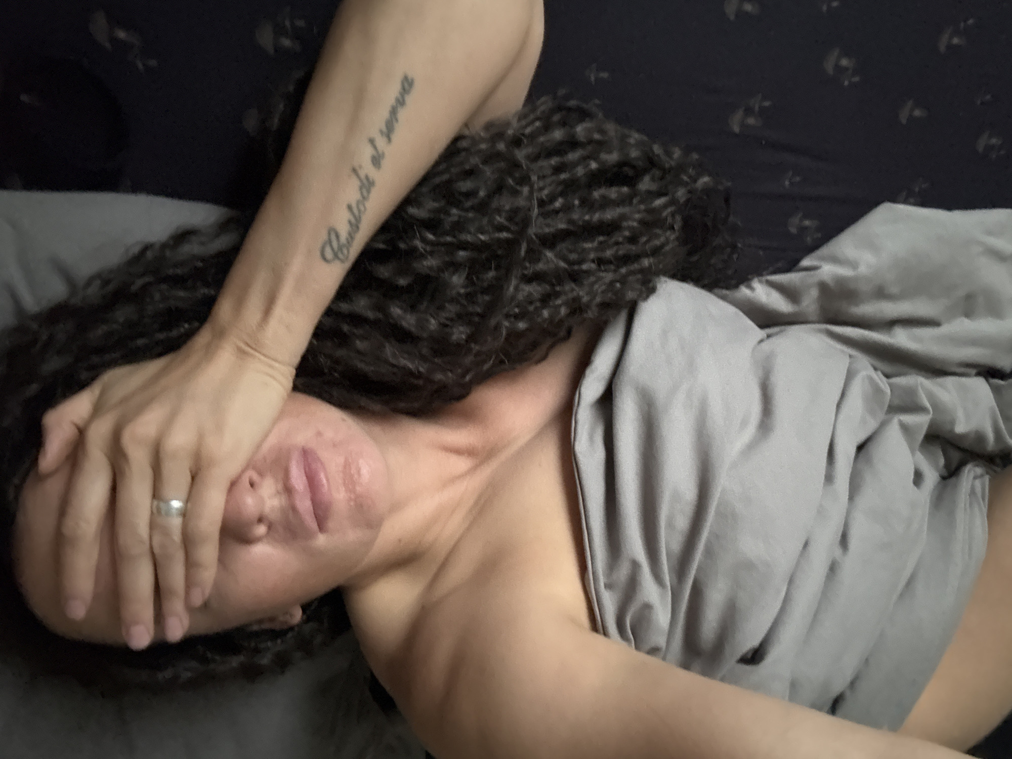 An overhead indoor shot captures a person lying on their side in bed. The individual has long, dark, curly hair that spreads across a dark pillow and bed. Their right arm is raised, with the hand covering their eyes and forehead, suggesting fatigue, discomfort, or a desire for privacy. A silver ring is visible on the ring finger of the hand covering the face. The person's face, partially visible, shows slightly parted lips and what appears to be some redness or flushing around the nose and mouth. A light grey blanket or sheet covers the lower part of their body. On the person's visible left forearm, a tattoo in script reads "Custodi et serva." The background behind the bed is dark with a subtle, indistinct pattern, indicating a dim-lit room. The scene depicts a quiet moment of rest or recuperation within a bedroom setting, possibly in Chaikovsky, Russia.