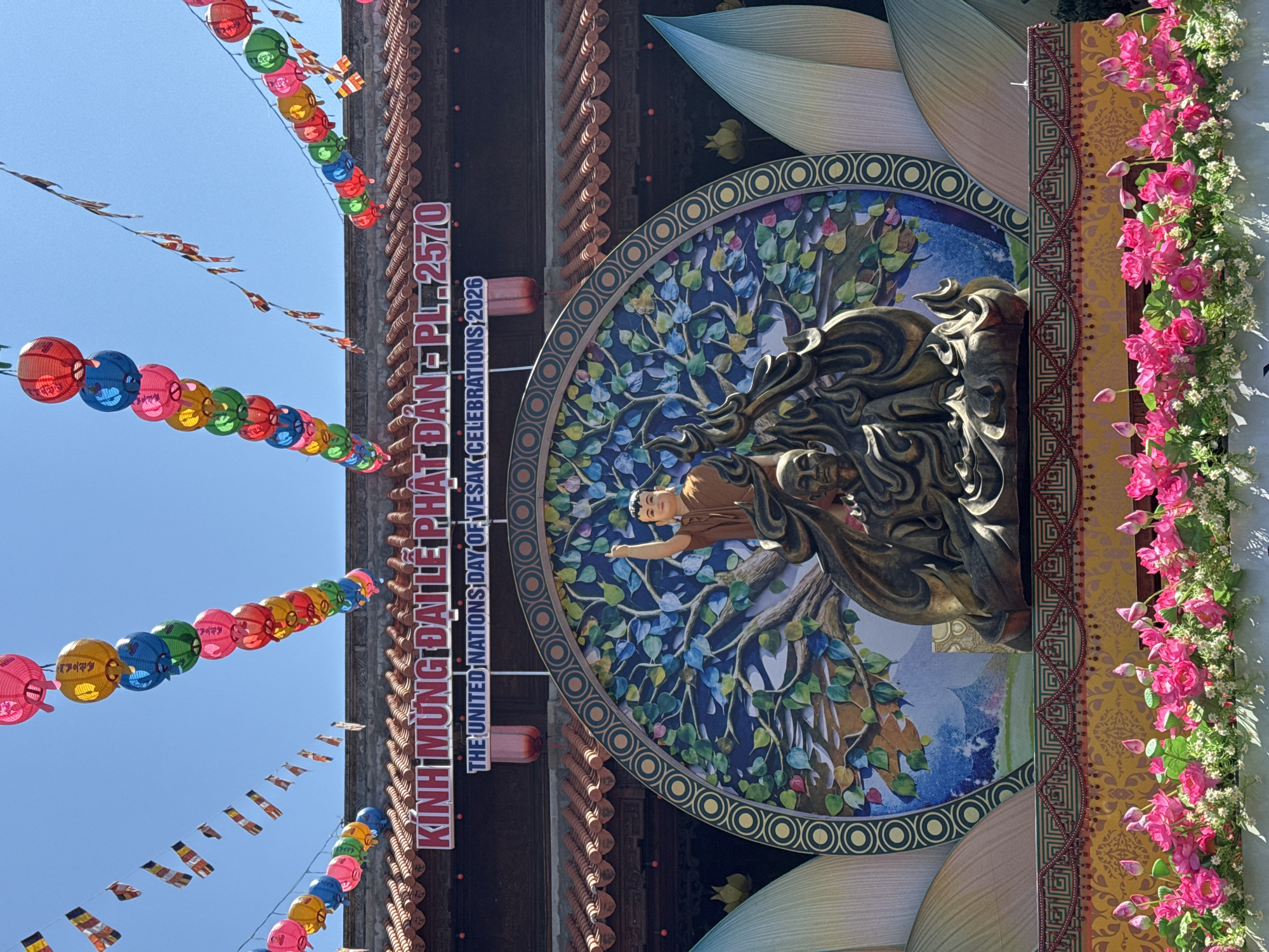 The image depicts a street decoration in Tay Nha Trang Ward, Vietnam. Colorful paper lanterns are strung across the street, along with small flags. A large sign in both Vietnamese and English, reads "KÍNH MỪNG ĐẠI LÊ PHẬT ĐẢN PL 2570" and "THE UNITED NATIONS DAY OF VESAK CELEBRATIONS, 2026." The sign features a circular graphic with a blue and green tree of life design behind a golden Buddha statue. To the right of the sign, there is an arrangement of pink flowers. The sky is visible in the background.