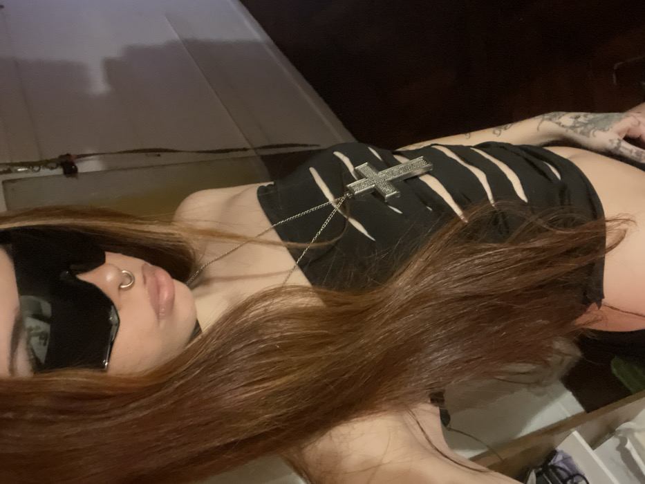 A female individual is depicted in an undetermined indoor location within the city of Unknown, Unknown. She is seen from the mid-chest upward, angled slightly to her left, with long, straight, reddish-brown hair cascading over her left shoulder and chest. Her eyes are obscured by large, dark, reflective shield-style sunglasses. She has a silver-colored septum piercing and light-colored, slightly parted lips. A black choker is visible on her neck. She wears a black strapless top featuring multiple horizontal cut-outs across the torso, exposing skin, and a long silver-colored chain necklace with a prominent, textured silver-colored crucifix pendant. Dark ink tattoos are partially visible on her left forearm. The background consists of a light-colored wall on the right with a distinct shadow, a dark floor section, and a partial white shelf or furniture item on the far left. The scene is dimly lit.