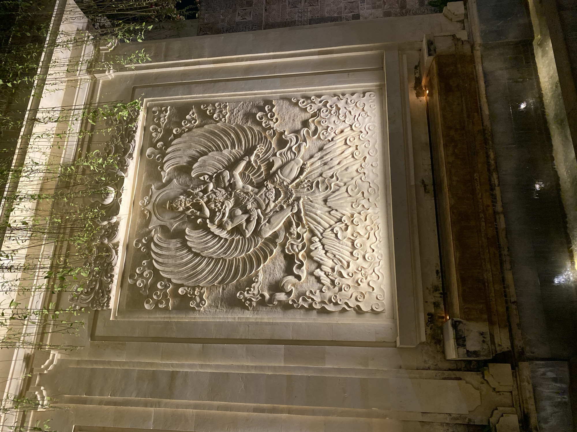 The image shows a stone relief carving of a mythological creature, possibly a phoenix or a Garuda, set into a wall at night. The carving is illuminated by artificial lights, creating dramatic shadows that highlight the intricate details of the creature's wings, feathers, and facial features. The creature appears to be in motion, with its wings spread wide and its body in a dynamic pose. Swirling cloud-like patterns surround the creature, adding to the sense of movement and fantasy.
The relief is framed by a decorative border, and the surrounding wall is made of large stone blocks. To the left of the carving, lush green vines with delicate leaves cascade down the wall, adding a touch of nature to the scene. To the right, a dark, wet surface, possibly a water feature or a wall that has been splashed, reflects the light. The overall setting suggests an outdoor space, perhaps a garden or courtyard of a temple or a luxurious establishment. The lighting indicates it is nighttime. There is no visible text in the image. The location context provided is Kuta Selatan, Indonesia, which aligns with the ornate and tropical aesthetic.