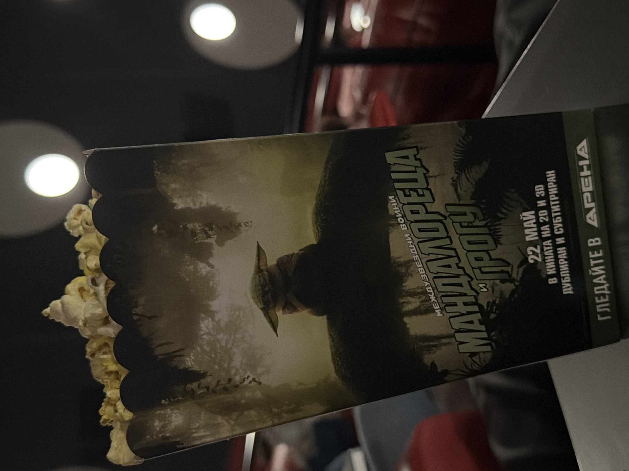 The image shows a popcorn box with a design related to the Star Wars series "The Mandalorian". The text on the box is in Bulgarian and translates to "The Mandalorian and Grogu" with the subtitle "Interstellar Wars." The text also includes "May 22," "In Cinema in 2D and 3D," "Dubbed and Subtitled," and "Watch in Arena." This indicates the photo was taken at the Arena cinema, where "The Mandalorian and Grogu" was shown, likely in Pleven, Bulgaria, given that Arena is a cinema chain with a location there.