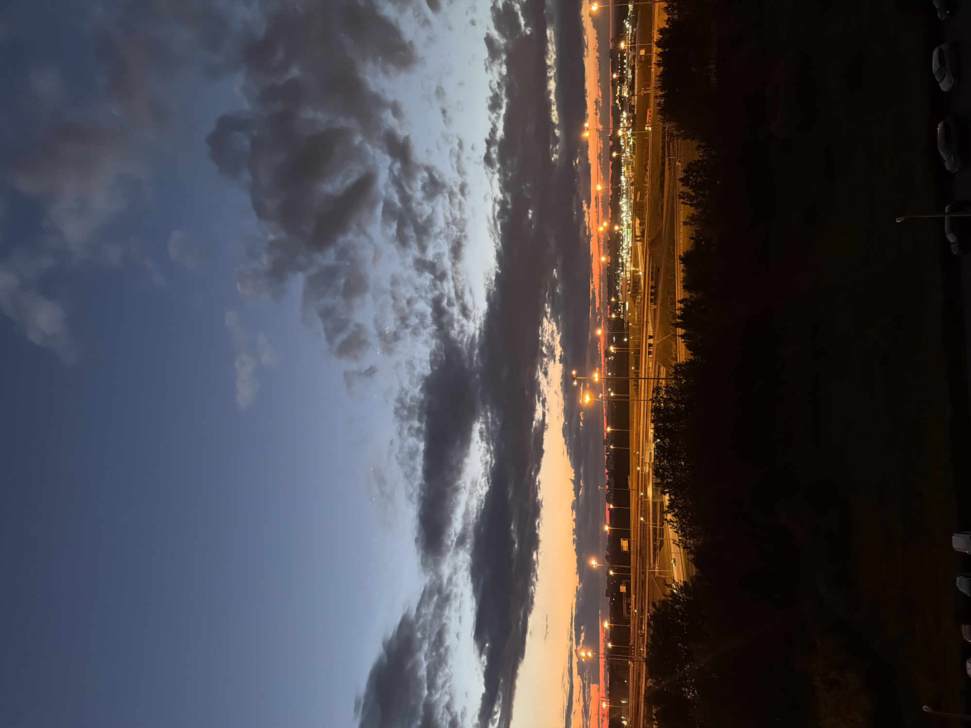 An eye-level outdoor shot captures a wide view of a city at dusk. The sky transitions from a deep blue at the top to a fiery orange and yellow near the horizon, indicating the setting sun. Dark, heavy clouds are scattered across the sky, adding depth and drama to the scene. Below the sky, a silhouetted line of trees forms the foreground, obscuring the ground. Beyond the trees, a network of elevated highways and roads is illuminated by numerous streetlights, casting a warm glow. In the distance, a cityscape is visible, with more lights dotting the urban landscape. The overall impression is one of a bustling city transitioning from day to night, with the artificial lights of the city contrasting with the natural beauty of the sunset and clouds.