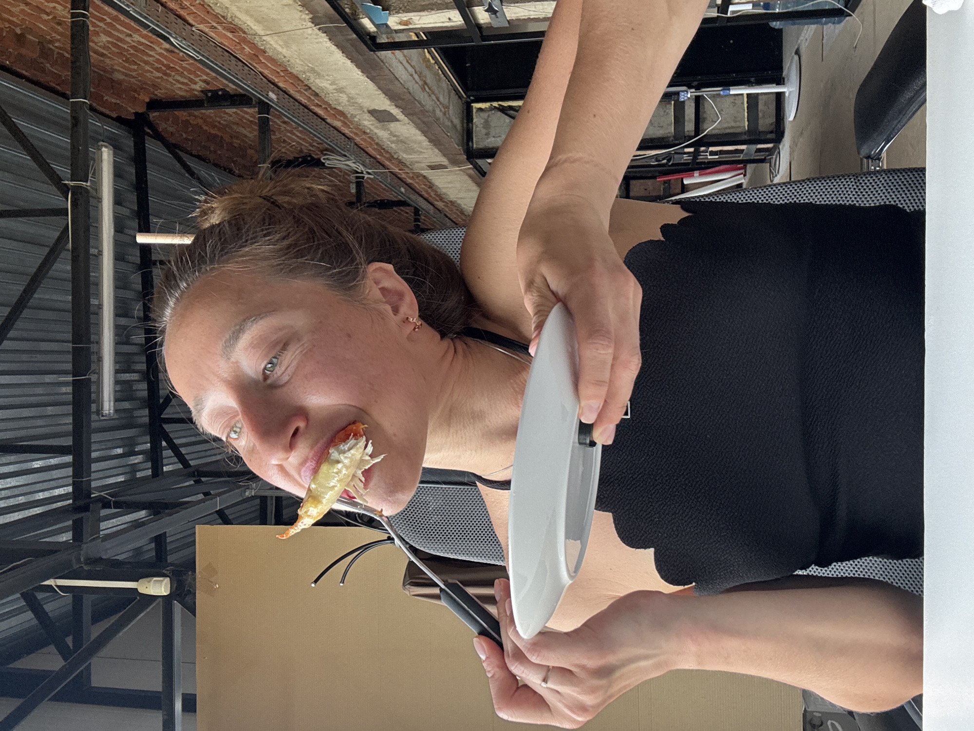 A woman with brown hair pulled back in a bun eats a shrimp with a fork and knife. She holds a white plate in her other hand. She wears a black shirt with scalloped edges at the neckline and what appears to be a black tank top underneath. She has pierced ears and wears a ring on her left ring finger. The background includes what appears to be a metal ceiling structure, lights, brickwork, and a cardboard surface. The location and city cannot be determined from the image.