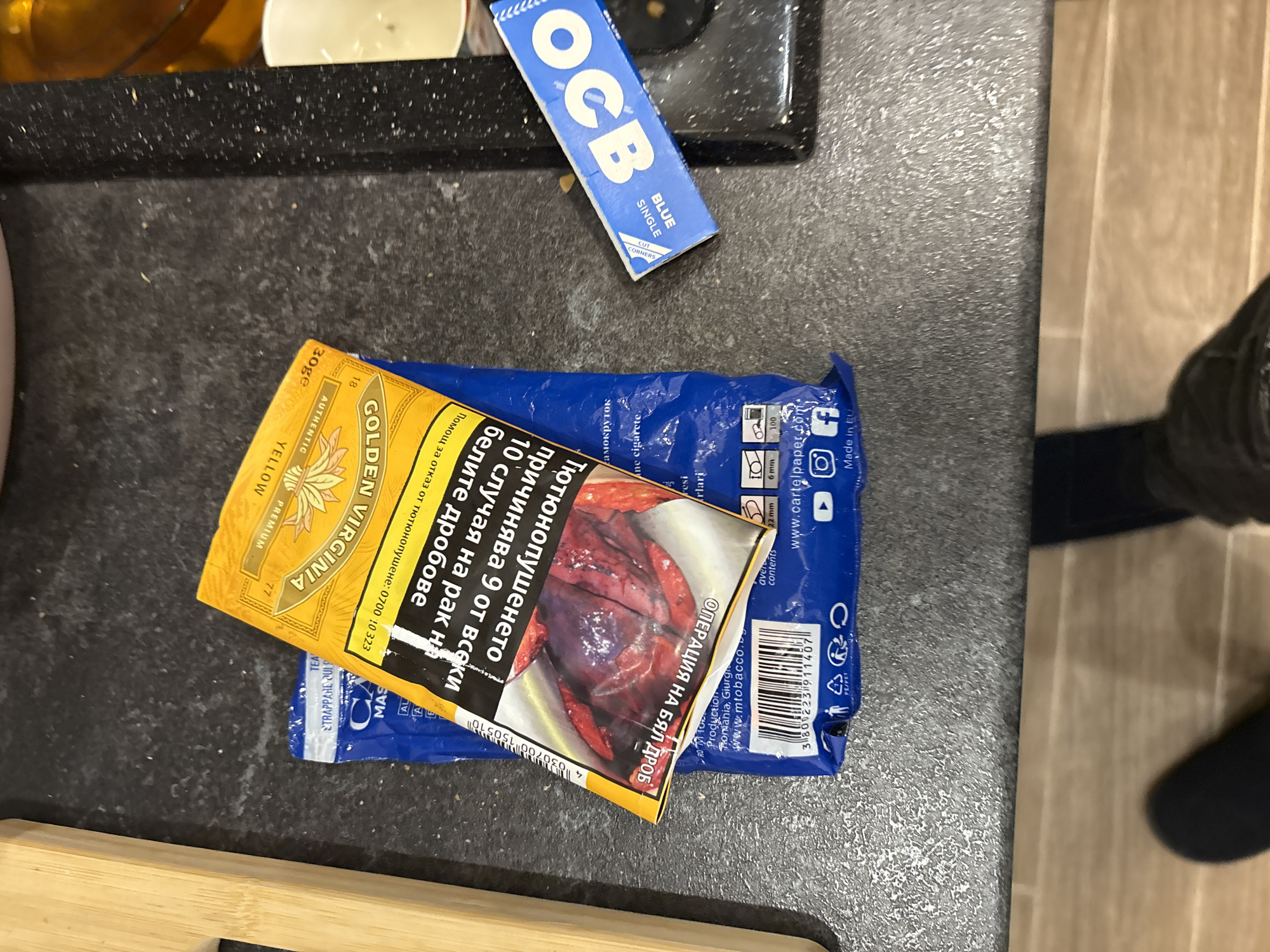 The image shows a countertop surface with various items. A yellow pouch of "Golden Virginia" tobacco is partially covering a blue package with the text "TEA", "MAS", and "TRAPPARE QUI P". The tobacco pouch features a graphic image of a lung, potentially damaged. Next to these is a blue pack of "OCB" rolling papers. A person's leg and footwear are visible in the background, suggesting someone is standing nearby. The countertop is a dark, speckled material. The presence of the tobacco products and rolling papers suggests smoking-related activities. There is no information to indicate that this is in Lovech, Bulgaria.