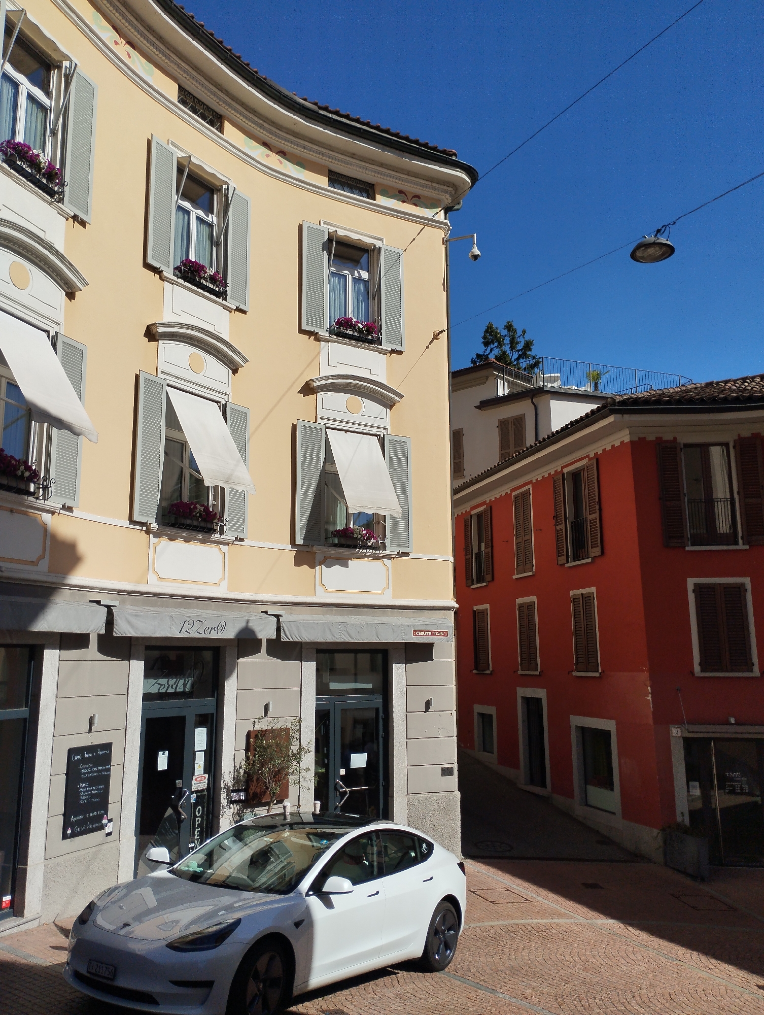 This image captures a sunlit street scene in Lugano, Switzerland, showcasing architectural details and urban life. The foreground features a white electric car, a Tesla Model 3, parked on a cobblestone street. Behind the car, a building with a yellow facade curves along the street. This building has multiple windows, some with open gray shutters and awnings, and flower boxes containing purple flowers. Below the windows, the building houses shops or cafes, with signage visible, including "12Zer()".
Adjacent to the yellow building is a vibrant orange building with dark wooden shutters on its windows. The street itself is paved with textured cobblestones. In the background, a clear blue sky is visible, with power lines and a lamppost spanning across it. The overall impression is of a charming European street with a modern element introduced by the electric car.