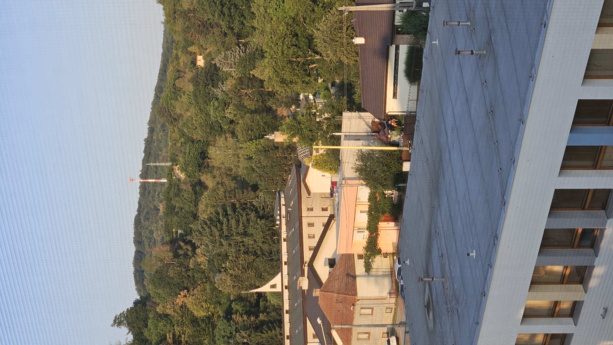 The image is an aerial view of a town with buildings and a densely wooded hill in the background. In the distance, a red and white striped telecommunication tower stands on the hill, partially obscured by trees. Several multi-story buildings with flat roofs and visible ventilation pipes are in the foreground and mid-ground. Some of these buildings appear to be institutional or industrial, with rows of windows. There are also smaller residential-style buildings with pitched roofs. Vehicles are visible parked on the street. The sky is clear and blue. The overall impression is a suburban or semi-urban environment adjacent to a natural, forested area. This scene is consistent with the southeastern part of Prešov, Slovakia, specifically an area that borders the Slanské vrchy mountain range.