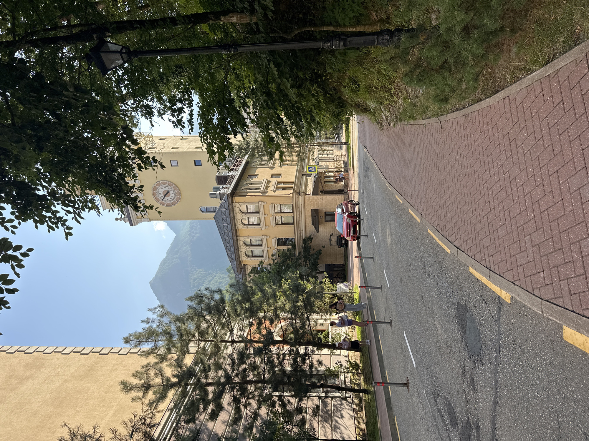 The image depicts a street scene in the city of Estosadok, Russia, under a clear blue sky. The street is paved with asphalt and bordered by a red brick sidewalk. Several street lights, marked by poles with red and white tops, line the road.
On the left side of the street, a tall, beige building with a textured facade and decorative elements stands prominently. In front of this building, several young people, appearing to be teenagers, are standing on the sidewalk, looking in different directions. Pine trees with dense foliage are planted along the sidewalk, casting shadows on the buildings and the ground.
Across the street, a yellow building with arched windows and ornate architectural details is visible. To the right of this building, a tall, yellow clock tower rises, featuring a large clock face with Roman numerals. A red car is parked on the street near the yellow building.
To the right of the sidewalk, a dense forest area with lush green foliage and tall trees dominates the landscape, creating a natural backdrop. The overall scene conveys a sense of a tranquil, picturesque urban environment integrated with nature.
