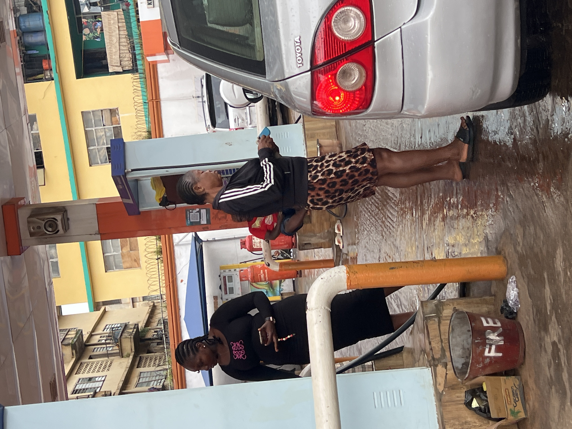 The image captures a street scene in Nkpor, Nigeria, likely at a gas station or a similar commercial area. Two women are the primary subjects, both dressed in casual attire. The woman in the foreground is wearing a black long-sleeved dress with a distinctive pink logo on the chest and holds a mobile phone. Her hair is styled in braids. The second woman, further back and slightly to the right, is wearing a black jacket with white stripes on the sleeves and a leopard-print skirt. She is also holding a mobile phone.
The setting appears to be outdoors, with a silver Toyota car prominently visible. The ground is wet, suggesting recent rain or damp conditions. In the background, buildings with yellow and other muted-colored facades are visible, some with barred windows, hinting at a dense urban environment. There are also elements of commerce, such as gas canisters and what looks like a fire bucket with "FIRE" written on it.
The overall mood seems to be ordinary, with the women engaged with their phones. The lighting suggests it could be daytime, possibly overcast due to the wet ground. The scene depicts a slice of everyday life in this Nigerian locality.
