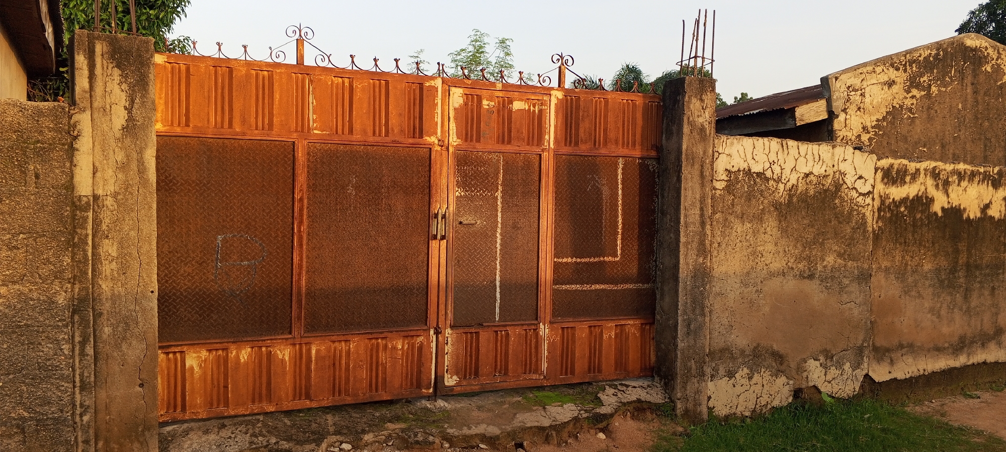 The image depicts a close-up view of a weathered, rusty, orange metal gate in Jalingo, Nigeria. The gate is made of vertical slats and features diamond-patterned metal panels in the upper sections. Decorative iron finials adorn the top of the gate. The gate is flanked by two rough, concrete pillars, one on the left and another on the right. The pillar on the right has exposed rebar at its top. To the right of the gate and pillar, a section of a cracked, aged concrete wall is visible, exhibiting signs of wear and staining. The ground in front of the gate is uneven, with patches of dirt, small stones, and some green grass. In the background, out of focus, are trees and a cloudy sky. The overall scene suggests an outdoor environment, possibly a residential or commercial property entrance, characterized by age and exposure to the elements.